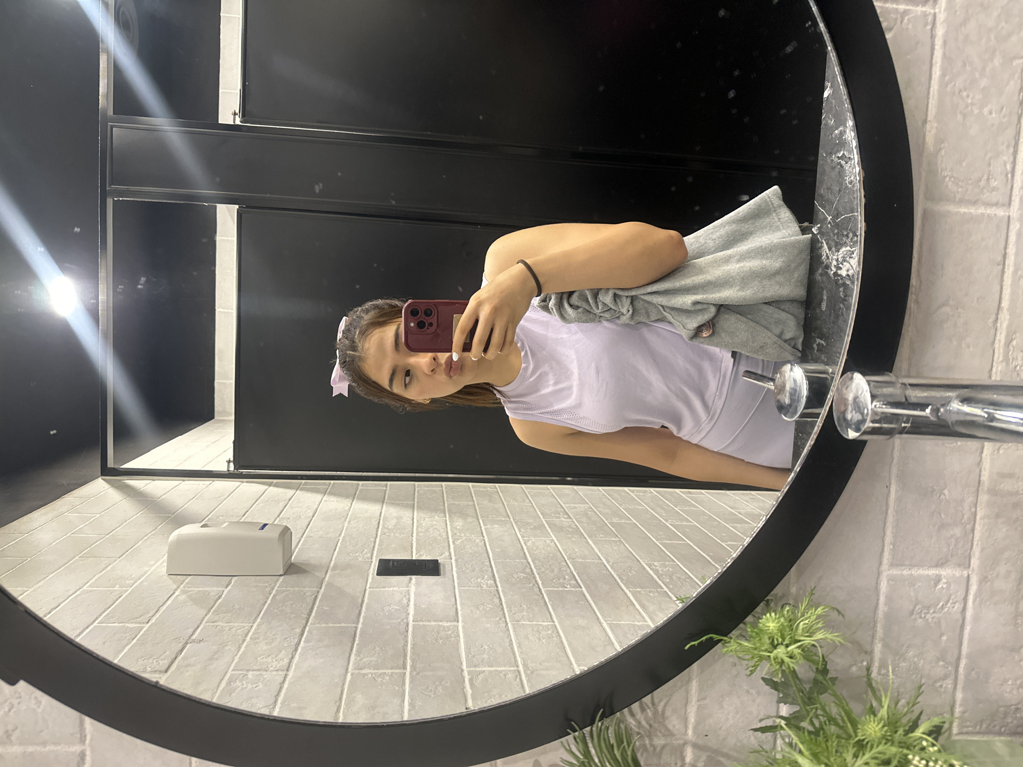 A woman is taking a selfie in a public restroom, likely in San Bernardino Tlaxcalancingo, Mexico. The woman is wearing a light purple athletic top and holding a red phone. She has a bow in her hair. The restroom has black stall doors, light gray tiled walls, and a round mirror. A soap dispenser is mounted on the wall near the sink. A gray garment is draped over the edge of the mirror. Green plants are in a vase near the sink.