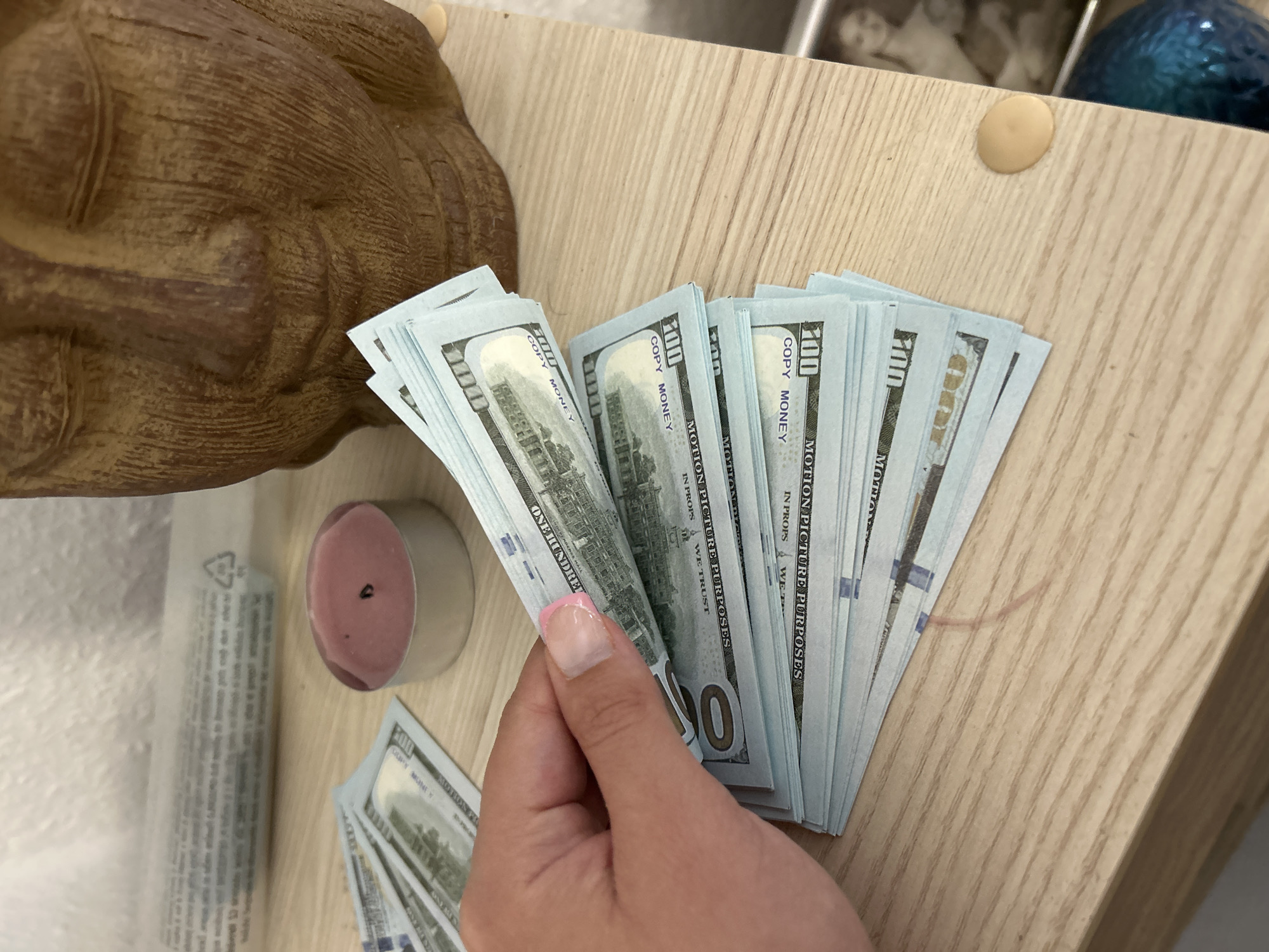 The image displays a close-up overhead view of a light-colored wooden surface. A fair-skinned hand, exhibiting manicured fingernails with pink tips, holds and fans out multiple stacks of prop U.S. dollar bills. Visible text on the bills includes "COPY MONEY" and "FOR MOTION PICTURE PURPOSES," with a clear "100" denomination, and they possess a distinct blue tint. To the right of the hand, a pink tea light candle sits in a silver metallic holder. In the upper right portion of the frame, a textured, brown sculpture resembling a Buddha's head with closed eyes is partially visible. The background includes a blurred translucent object with faint text. On the wooden surface, several light-colored circular caps, likely screw covers, are present, along with a thin, reddish-brown fiber. In the lower right corner, a portion of a blue, textured spherical object and a dark-framed image containing a person's likeness are visible.