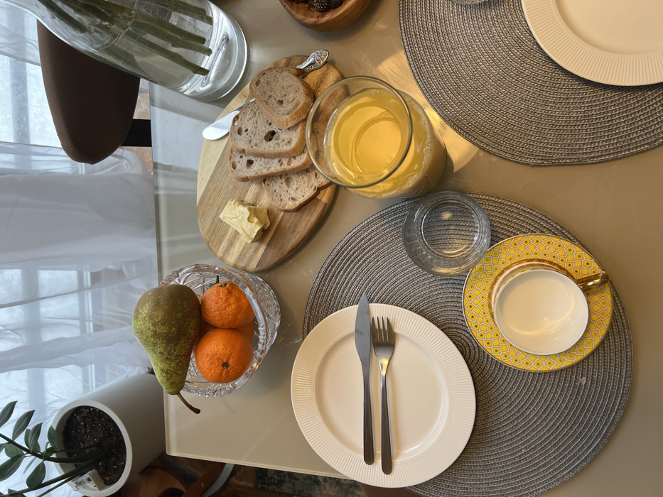 The image depicts an overhead view of a dining table set for a meal in a residential interior in Khimki, Russia. The light-colored table, possibly glass, reflects ambient light.
In the upper-left, a white cylindrical planter containing a green leafy plant is partially visible. Centrally, a crystal bowl holds one green and brown pear and three orange tangerines. To the right of the fruit bowl, a two-toned wooden cutting board presents four slices of bread, a knob of butter, and a small silver-colored butter knife. Further right, a clear glass vase with green stems is partially visible, along with a portion of a brown upholstered chair back. A small wooden bowl containing dark berries is situated at the far right edge of the table.
Two round, woven grey placemats define place settings. The left placemat displays a white dinner plate with ribbed edges, topped with a silver-colored knife and fork. Below this plate, a yellow-patterned saucer with a white teacup (featuring a yellow and gold-accented handle) is present. Adjacent to the teacup and saucer, on the same placemat, is an empty clear glass tumbler. To the right, a tall glass filled approximately two-thirds with orange juice is positioned. Another partial placemat with a white plate is visible in the lower-right corner.
The background features a large window obscured by sheer white curtains, providing diffused natural light to the scene. No individuals are present.