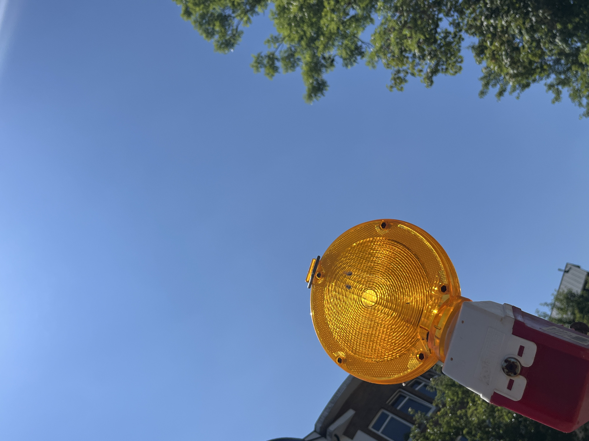 An orange traffic light is positioned in the lower-left quadrant of the frame, angled upwards against a clear blue sky. The light has a textured, circular lens with concentric grooves. It is mounted on a red and white housing, which appears to be a battery pack or a power source. The background shows a building on the left, with windows visible, and lush green trees on the right, their foliage blurred. The image was taken in Saint Vith, Belgium.