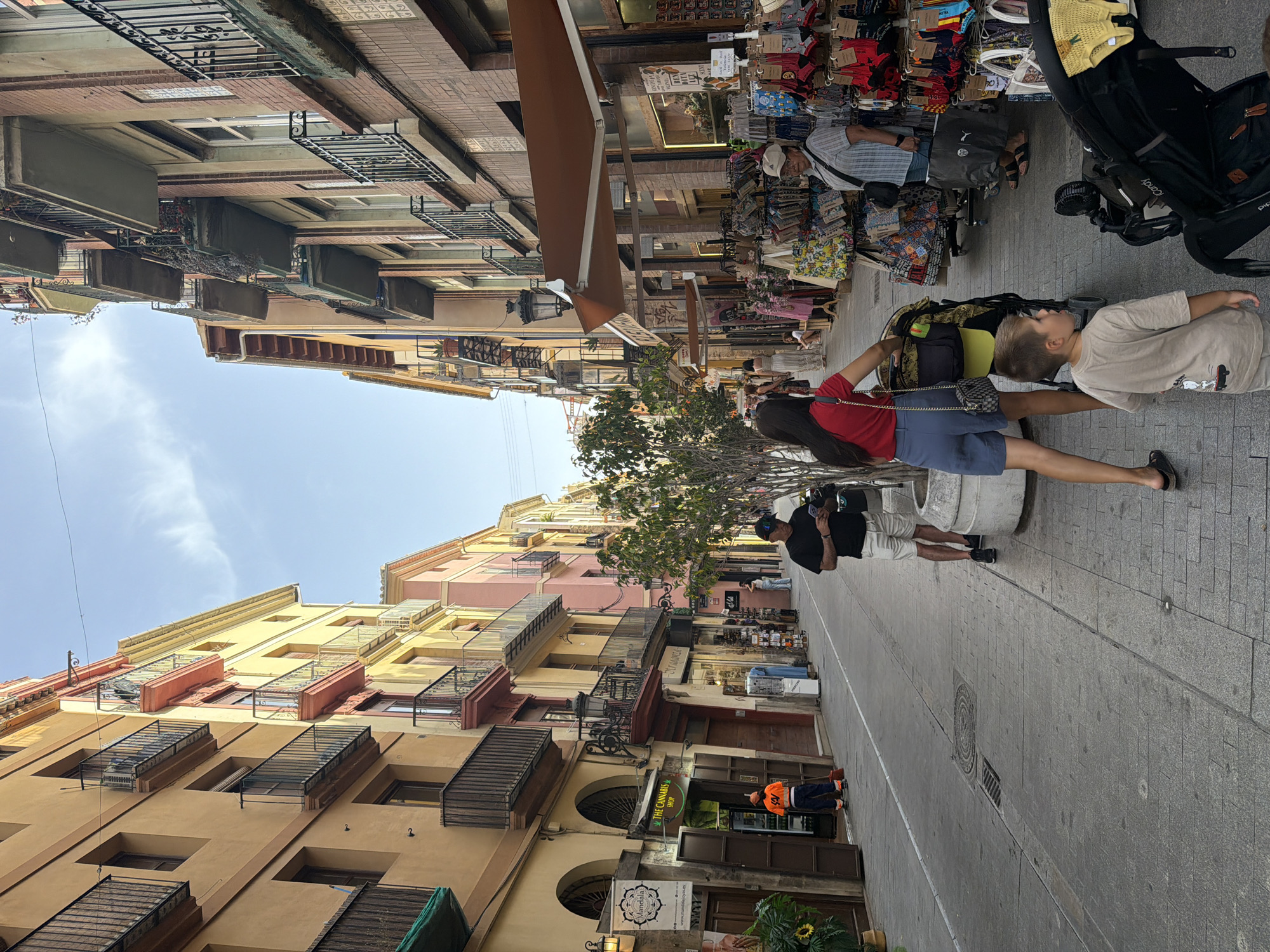 The image captures a street scene in a city with buildings lining both sides of a pedestrian walkway. The buildings are multi-story with balconies and windows. On the left side, there are shops with awnings and displays. One shop is labeled "THE CANNABIS SHOP". A person in an orange vest is sweeping the sidewalk in front of a shop. On the right side, there are more shops and market stalls displaying various goods, including clothing and bags. Several people are walking along the street. In the foreground, a woman in a red top and blue shorts is pushing a stroller. A young boy in a beige t-shirt is walking ahead of the stroller. Another person in a black t-shirt and white shorts is looking at their phone. The sky is visible at the top of the image, with a few clouds. The ground is paved with stone tiles. The overall scene depicts a lively urban environment with commercial activity and pedestrians.