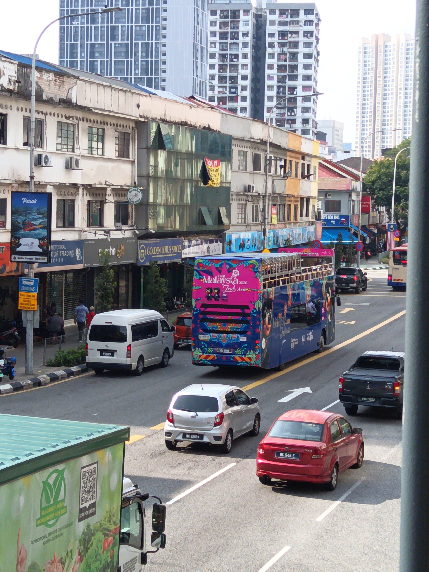 The image depicts a street scene in Kuala Lumpur, Malaysia, likely in the vicinity of Jalan Bukit Bintang or a similarly bustling commercial area. Tall, modern skyscrapers with numerous windows dominate the background, contrasting with the older, two-to-three-story buildings lining the street. These older buildings feature shops with awnings and signage, some appearing a bit worn.
In the foreground, a light green delivery truck is partially visible on the left, featuring a QR code and text in Malay. A white minivan is driving on the road, followed by a silver hatchback. A bright red sedan is in the adjacent lane. Further down the road, a colorful double-decker bus with "Visit Malaysia 2020" branding is prominent. A dark pickup truck is also on the road.
Pedestrians are visible on the sidewalk near the older buildings, with some sitting on a bench and others walking. Street lamps are positioned along the sidewalk. The road is paved and marked with white lane dividers and arrows. The overall impression is of a busy urban environment with a mix of old and new architecture, traffic, and commercial activity.