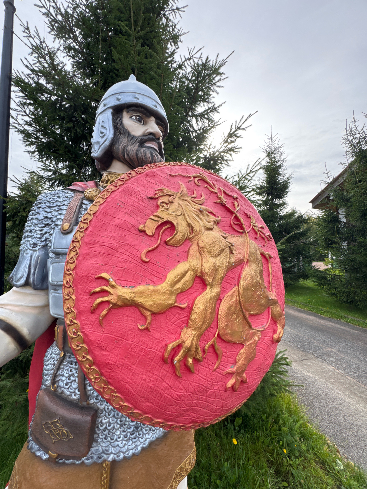 A life-sized statue of a warrior is depicted in the foreground, positioned outdoors in front of evergreen trees and a paved path. The warrior is wearing a silver helmet, chain mail, and a red cape. He holds a large, circular red shield adorned with a golden relief of a mythical beast. A brown leather pouch is attached to his belt. The background shows a cloudy sky, a portion of a building with a white facade and a brown roof, and the green foliage of trees and grass. The statue appears to be a decorative element, possibly at an entrance or along a pathway. The scene is set in Peterhof, Russia.