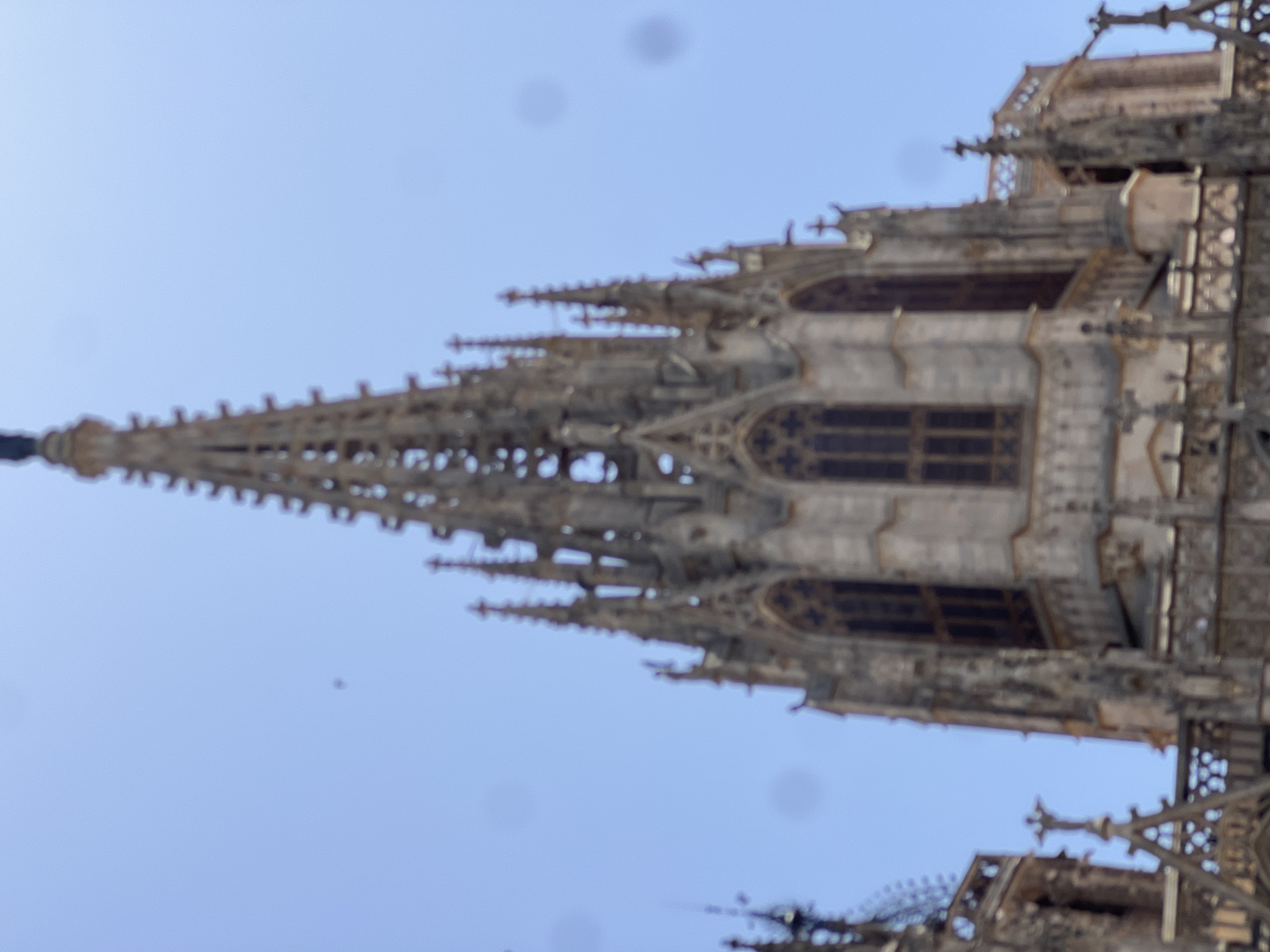 The image is a blurred, low-angle shot of the Gothic architecture of the Barcelona Cathedral in Barcelona, Spain, under a clear blue sky. The focus is on the intricate spires, stained-glass windows, and stone detailing of the cathedral's facade. There are several circular, dark smudges on the image, likely dust or artifacts on the lens. The main spired structure dominates the center of the frame, with smaller spires and architectural elements visible on the left and right. The sky is a bright, consistent blue, with no clouds.