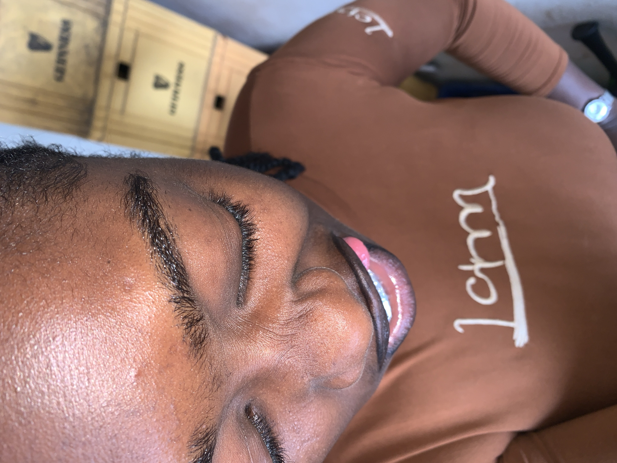 An overhead photograph captures a person with dark brown skin, located in New Stadium, Nigeria. The individual is looking downwards, with eyes closed or heavily lidded. Dark eyelashes and eyebrows are prominent. The nose shows distinct wrinkles and folds, consistent with a smiling expression. The mouth is open, revealing white teeth and a pink tongue slightly extended. The person is wearing a fitted, long-sleeved, rust-colored or terracotta-toned shirt. White script text is visible on the chest, reading "I am", with additional partial text on the right sleeve. A silver-colored watch is present on the left wrist. In the blurred background, two light-colored, possibly wooden or cardboard boxes are stacked. Each box features a dark label with white text and a harp logo, which is identifiable as "GUINNESS".