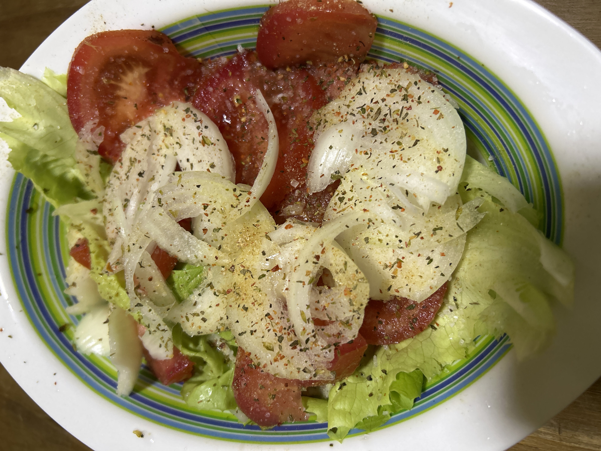 A close-up, overhead shot showcases a vibrant salad in a colorful bowl. The salad consists of fresh green lettuce, sliced red tomatoes, and thinly sliced white onions, all sprinkled with a savory seasoning blend. The bowl itself features a striking pattern of green, blue, and white stripes. The background is a blurred wooden surface. The image is set in an unspecified location within the city of Buenos Aires, Argentina.