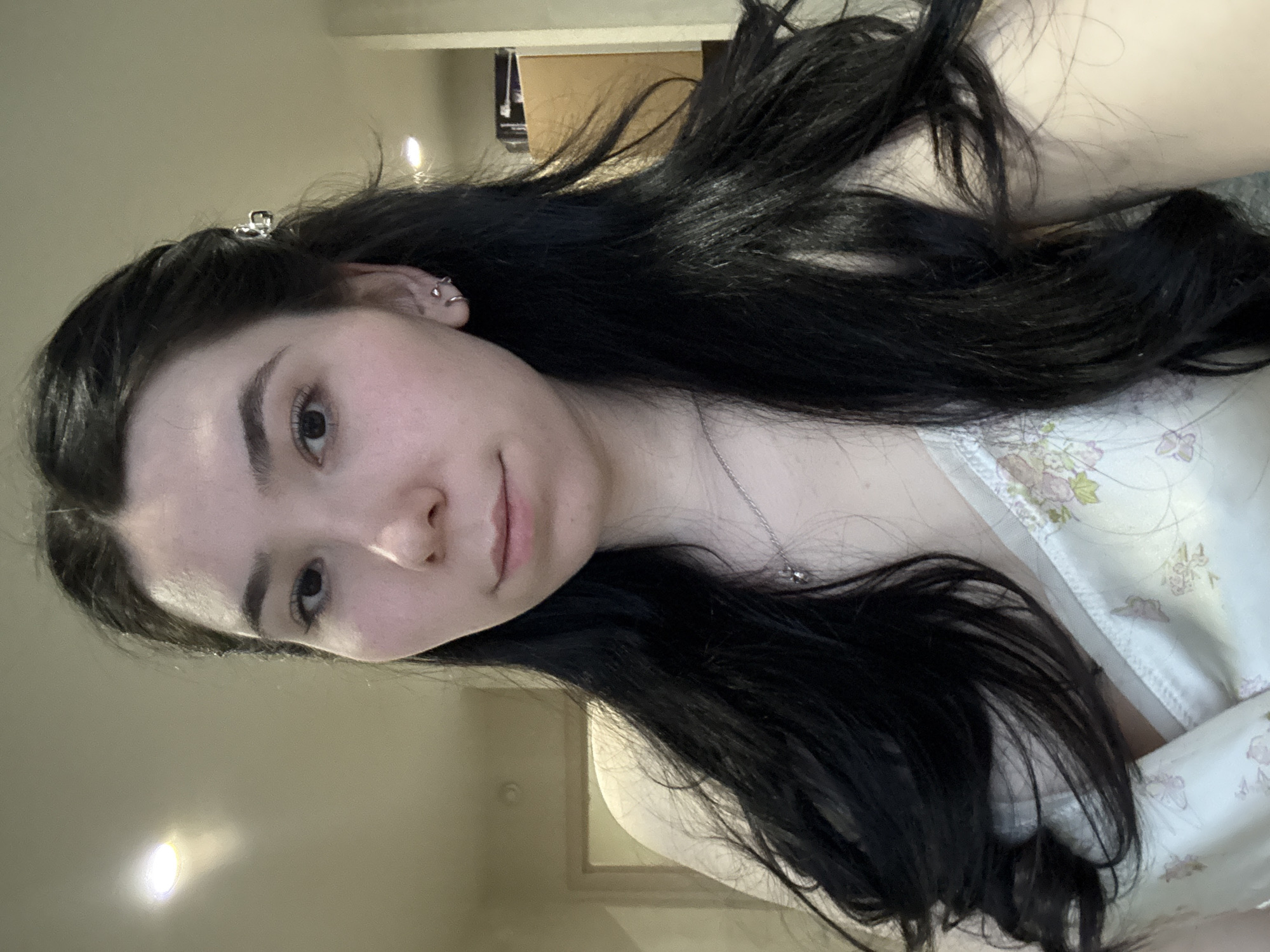 A person with long, dark hair is in a brightly lit interior. The person appears to be facing the camera, with their head slightly tilted. The person has fair skin, dark eyebrows, and a small silver hair clip in their hair. A silver necklace adorns their neck, and they are wearing a floral-patterned top. The background is blurry but shows a doorway and beige walls. There is no evidence to suggest the location is Izhevsk, Russia.