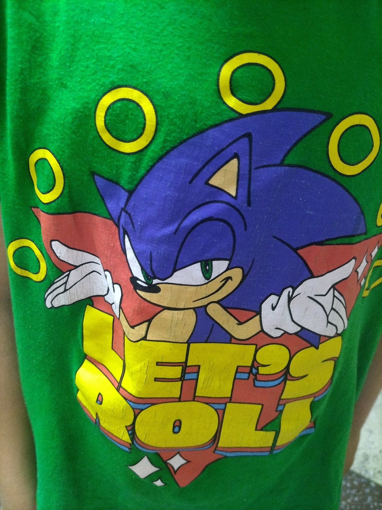 The image shows a close-up of a green t-shirt with a graphic of Sonic the Hedgehog. Sonic is depicted in his classic blue color, with green eyes and a red cape. He is pointing with his right hand, and the text "LET'S ROLL" is written in large yellow letters below him. The graphic also includes several yellow rings scattered around Sonic, a common element in the Sonic video games. The t-shirt is being worn by a person, whose arm is partially visible on the left side of the image. The background is out of focus and appears to be an indoor environment. There is no information in the image to suggest any location in the city of Hrodna, Belarus.