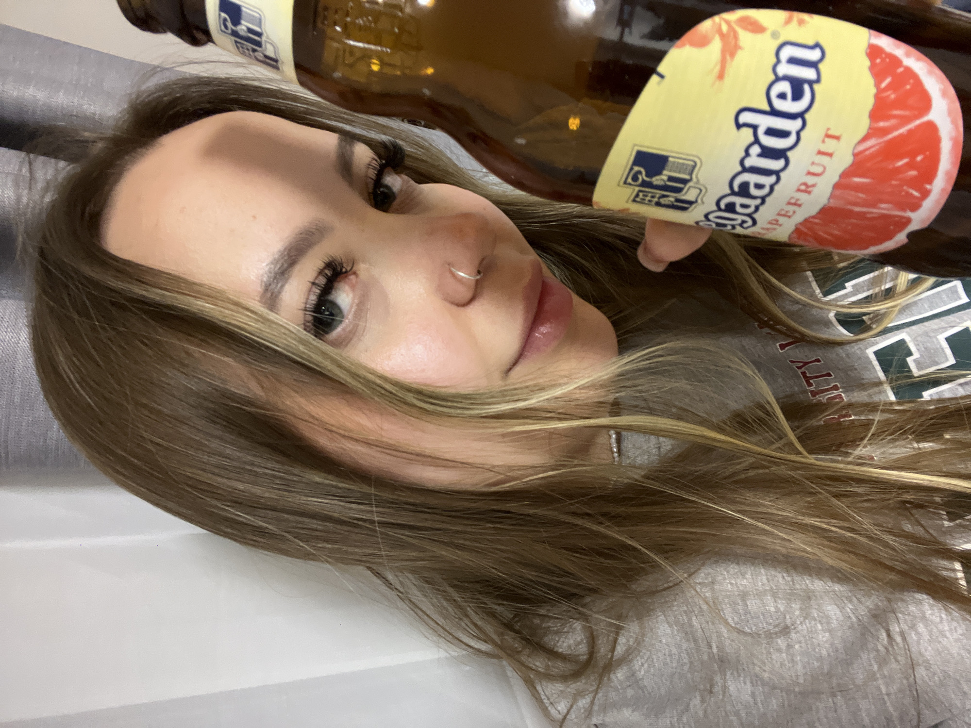 This portrait-oriented image, rotated clockwise, features a young woman lying down, looking towards the camera with a relaxed expression and a soft smile. She has long, light brown hair with blonde highlights, which is spread around her. Her face is visible from the forehead to the chin, revealing well-defined eyebrows, prominent false eyelashes, and a small silver hoop nose piercing. Her lips are slightly parted. She is wearing a grey top, possibly a sweatshirt, with some dark text partially visible on the right side, resembling '...ITY' from a word like 'UNIVERSITY'. A brown glass bottle, labeled 'Hoegaarden Grapefruit', is held above her head or rests near her. The label clearly shows the brand name 'Hoegaarden' and the flavor 'GRAPEFRUIT', along with a logo featuring a building. The background is simple, consisting of a grey fabric texture behind her head and a plain white surface to her left. The overall scene conveys a casual and relaxed indoor moment. The given location context for this photo is Krasnodar, Russia.