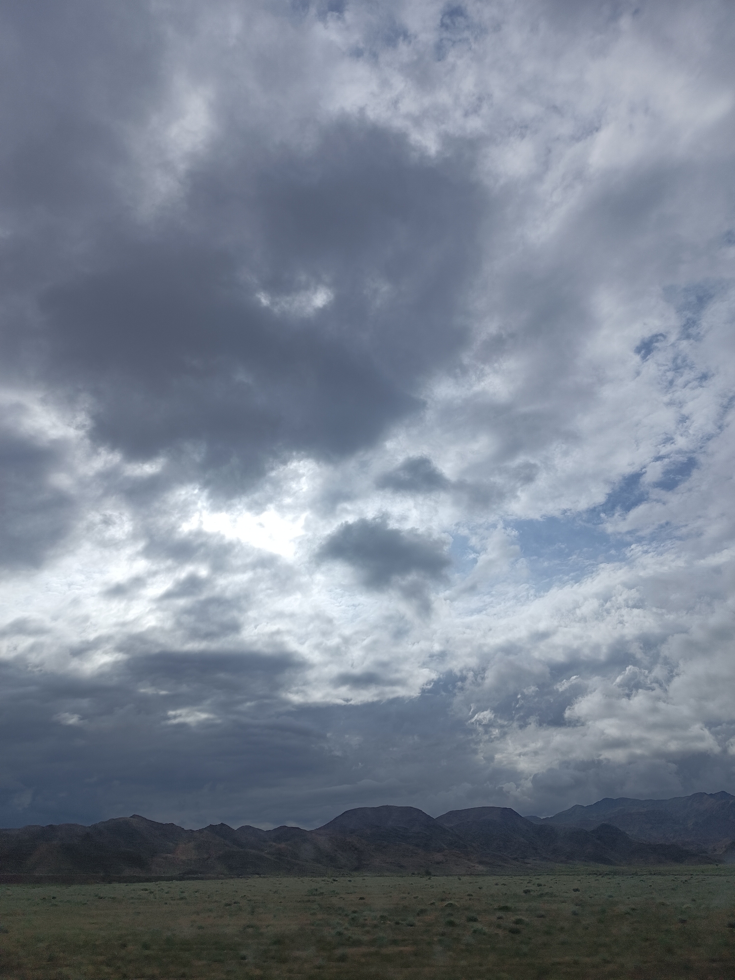 The image depicts a wide, horizontal view of a landscape under a dramatic, cloudy sky. The foreground consists of a flat, grassy plain with sparse vegetation, receding towards a range of dark, undulating hills in the mid-ground. These hills, appearing barren and rugged, stretch across the entire width of the image. Above the hills, the sky is filled with heavy, grey clouds, with pockets of brighter light breaking through in places, suggesting the presence of the sun. There are no people, animals, or man-made structures visible in the scene. The overall impression is one of a vast, natural environment, possibly a steppe or mountainous region, under overcast or stormy weather conditions. The location is identified as Sasyk-Bulak, Kyrgyzstan.