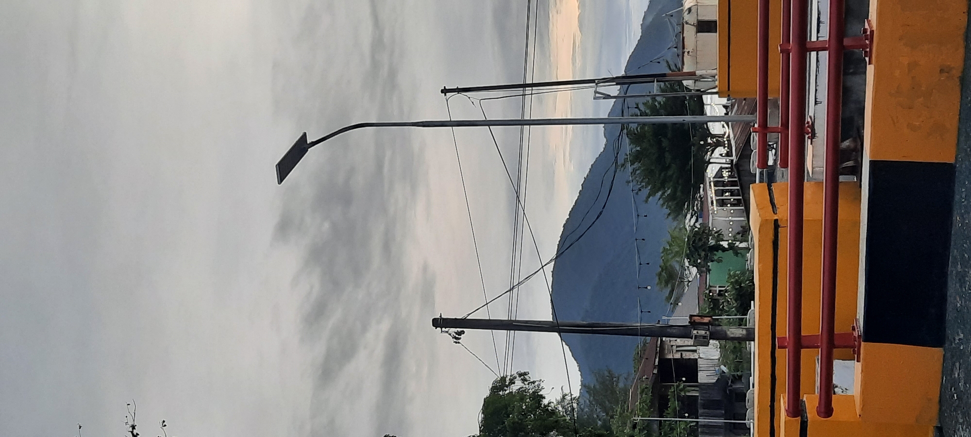 This image captures a view in Banda Aceh, Indonesia, likely near the coastline or a body of water, given the presence of a hill that could be a coastal mountain range. The sky is overcast with gray clouds, indicating a potentially humid or rainy day.
In the foreground, there are safety barriers, painted in bright yellow and black, with red railings. These barriers appear to be along a road or walkway, possibly a bridge or an elevated structure.
Further back, several utility poles with visible electrical wires stretch across the frame, contributing to the urban or semi-urban landscape. One prominent modern street lamp with a solar panel on top stands tall to the right of the center.
Behind the utility poles and barriers, a lush green hill or small mountain dominates the background, its slopes covered in vegetation. The lower slopes of the hill show signs of human settlement with scattered buildings, some with corrugated metal roofs, and trees. String lights are visible between some of the buildings, suggesting a commercial or recreational area.
The overall scene suggests a transition zone between an urban environment and the natural landscape, characteristic of coastal cities in Indonesia. The time of day appears to be either early morning or late afternoon, given the soft, diffused light. There are no visible people or animals in the frame.