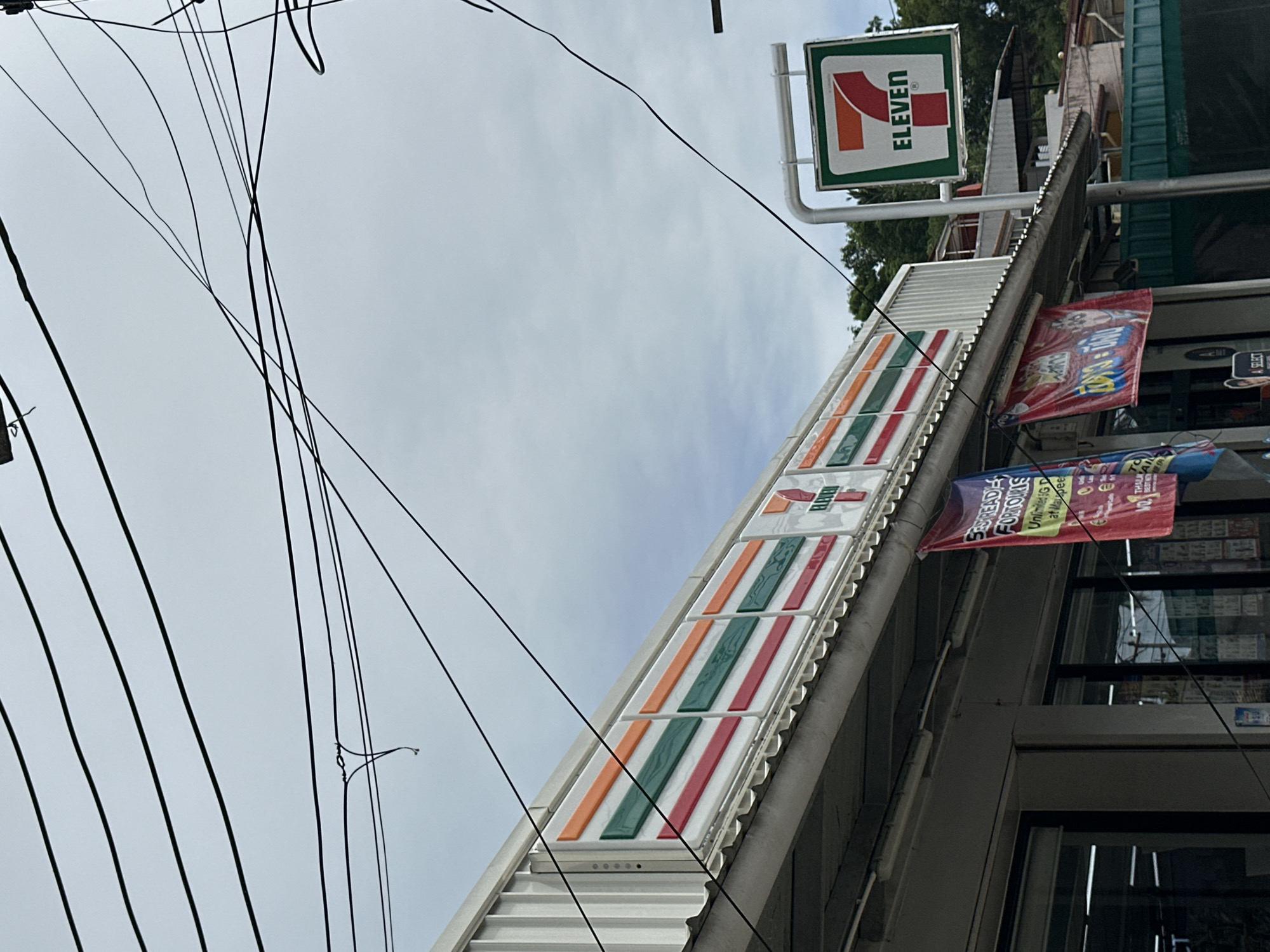 The image shows a 7-Eleven store in Karon, Thailand. The store has a white corrugated metal exterior with the iconic 7-Eleven logo visible on a signpost. There are also colorful banners hanging outside the store, advertising various promotions. The sky is overcast, and there are several power lines running across the frame. The scene appears to be a typical street view of a convenience store in a tropical location.