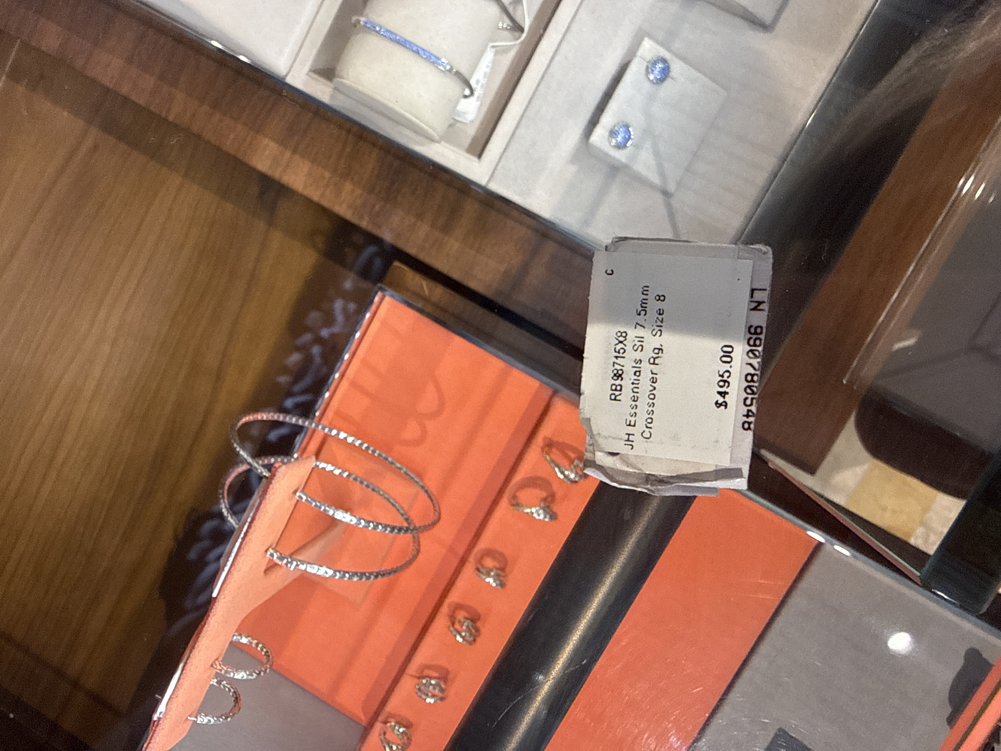 An overhead shot captures a display case containing various jewelry items. In the foreground, an orange card holds a selection of silver hoop earrings, some intricately patterned and others simple bands. To the right, a display box exhibits a delicate blue beaded bracelet and a pair of circular blue stud earrings. A price tag, partially obscured, indicates "$495.00" and lists "JH Essentials Sil 7.5mm Crossover Rg. Size 8". The background reveals a polished wooden surface, suggesting a retail environment. The scene is clean and well-lit, emphasizing the merchandise. No people are visible, and the location is a jewelry store, likely in the city of Kuta, Indonesia, based on the context.