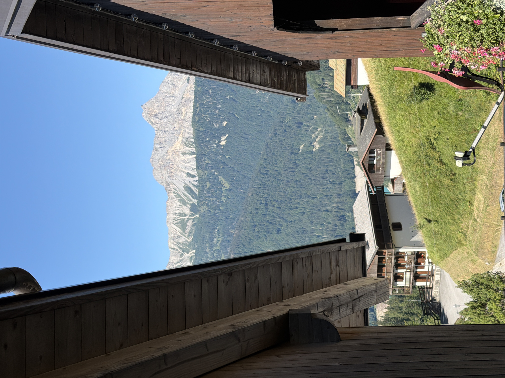 The image shows a view of a mountainous landscape with some buildings in the foreground, likely in Sauris/Zahre, Italy. The perspective is from between two wooden structures, possibly balconies or eaves, which frame the scene. The sky is clear and blue.
In the mid-ground, there are several buildings with traditional alpine architecture, featuring wooden balconies and pitched roofs. Some of these buildings are situated on a grassy hillside. A satellite dish is visible in the lower right corner, next to a bush with pink flowers.
The background is dominated by a large, rugged mountain range with rocky peaks and slopes covered in dense green forest. The sunlight suggests it is daytime, and the overall impression is one of a clear, sunny day in a mountainous region. No people are visible in the image.