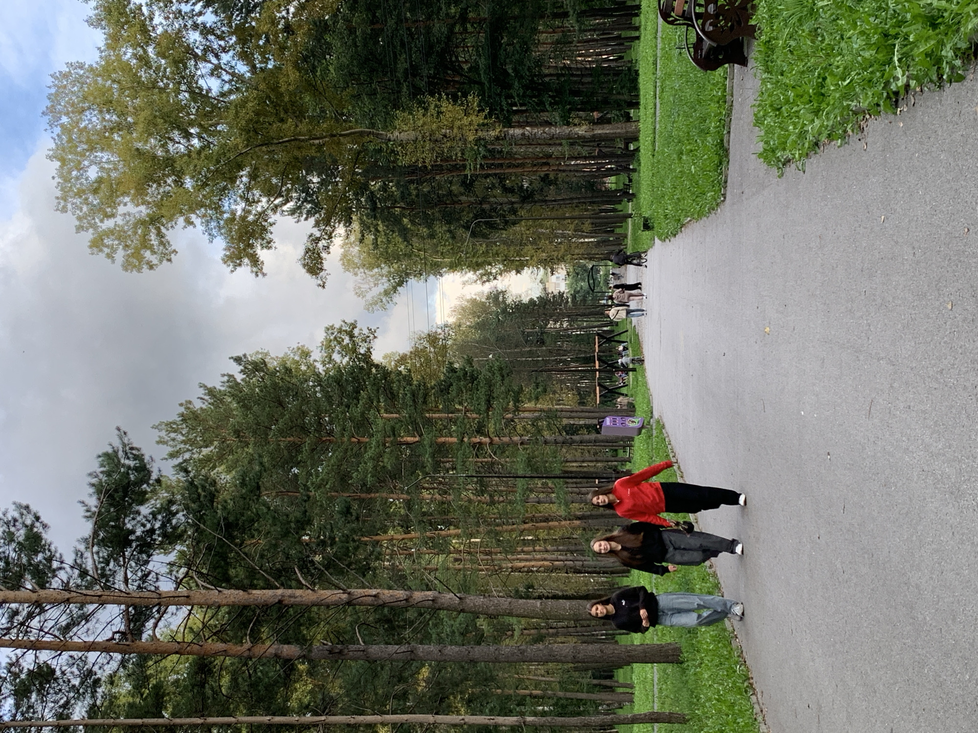 A paved pathway through a park lined with tall pine trees and green grass runs towards a distant building. Three young women walk along the path, two in casual attire and one in a red jacket. Other park-goers and children can be seen in the background, some on bicycles. The sky is overcast with patches of blue. This scene is typical of a public park in Prokopyevsk, Russia, a city known for its industrial heritage and surrounding forests.