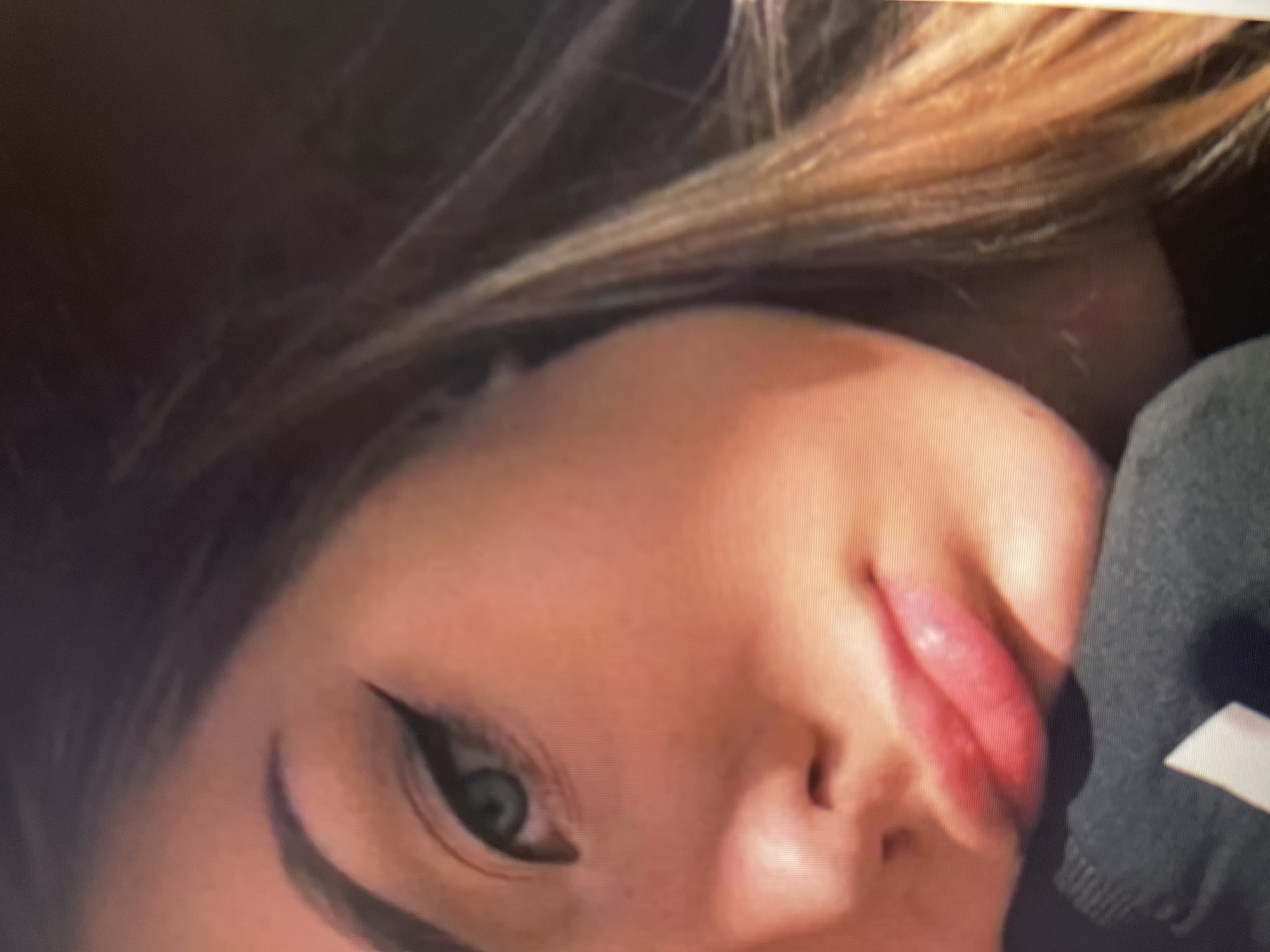 The image shows a close-up portrait of a young woman. She has light skin with a few freckles and blue-green eyes. Her eyebrows are dark and arched, and she is wearing winged eyeliner. Her lips are full and appear to be colored with a lip gloss. Her hair is long and light brown, with some lighter streaks, and is partially visible on the right side of the frame. She is wearing a dark-colored garment, possibly a sweatshirt or jacket, around her neck and shoulder. The lighting suggests she is in an indoor setting, with a light source creating a strong highlight on her cheek. The background is dark and out of focus, with some subtle highlights that could indicate reflections or a textured surface. There is no visible indication of the city of Qarağandı city administration, Kazakhstan.