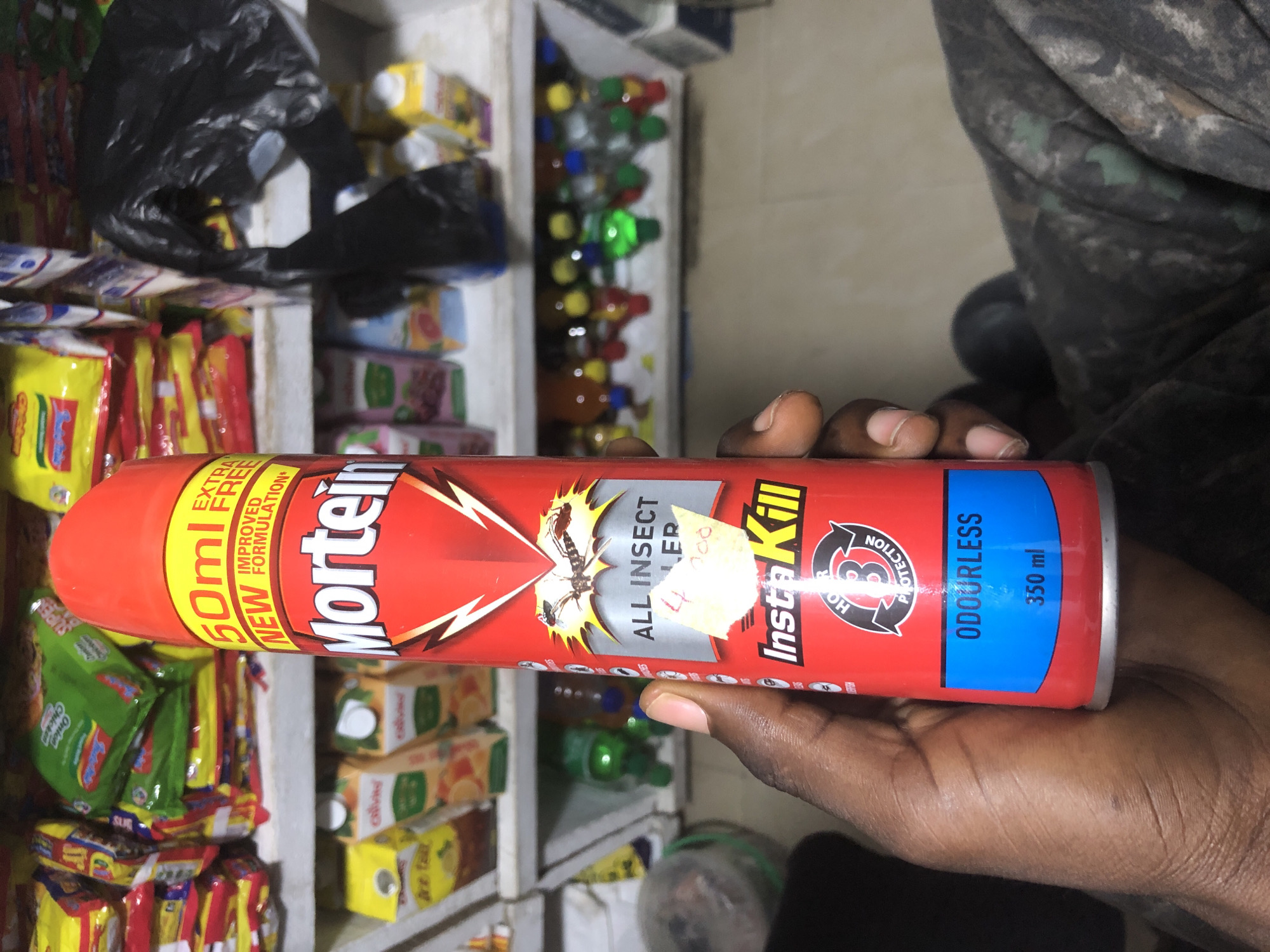 The image shows a hand holding a red aerosol can of Mortein InstaKill All Insect Killer. The can is approximately 350 ml and is labeled as odorless. The background consists of shelves stocked with various consumer goods, including snacks, juices, and small bottles. The setting appears to be a small retail store or a stall in a market in Ado, Nigeria, judging by the packaging of some products which are common in that region. A price tag of 4000 is visible on the can.
