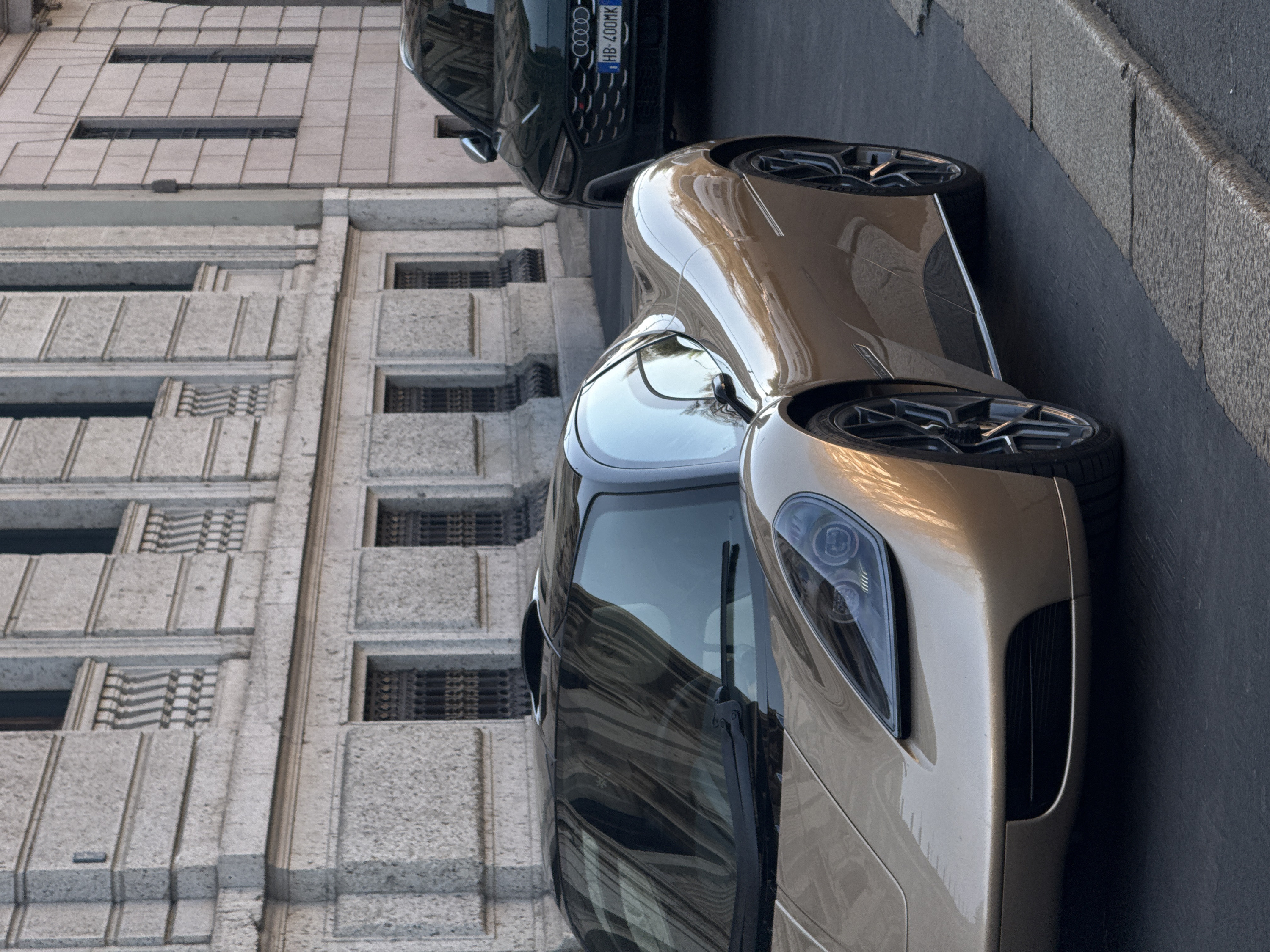 The image depicts a street scene in an affluent, historic district of Milan, Italy. In the foreground, a golden-bronze Gordon Murray T.50 hypercar is positioned at an angle, facing forward and slightly to the left, occupying the left side of the frame. The vehicle features a sleek, aerodynamic body, a black roof panel, distinctive LED headlights, and dark multi-spoke alloy wheels. Behind the hypercar, partially obscured, is a dark green Audi RS SUV, also stationary. Its front grille, four-ring emblem, and an "RS" badge are visible, along with a Hungarian license plate displaying "HB-400MK." The background consists of a multi-story classical-style building with a light-colored, textured stone or stucco facade. The building exhibits architectural details such as horizontal banding, rectangular block sections, and tall, narrow windows. Several windows on the upper floors are adorned with decorative dark iron grilles and balustrades beneath. The street surface is dark asphalt, bordered by a rough-hewn granite curbstone in the lower right. No individuals are visible, and no discernible actions or interactions are taking place.