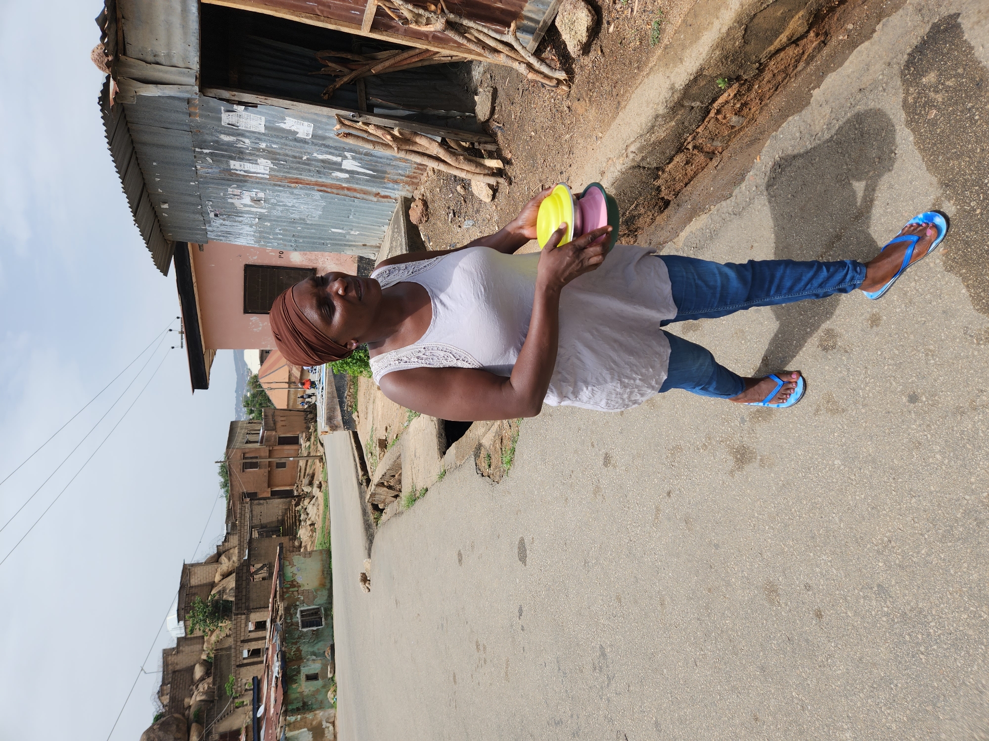 A woman stands on the side of a paved road in Jos, Nigeria. She wears a white top, blue jeans, and blue flip-flops. A brown headscarf covers her hair. She holds a stack of colorful bowls in her hands. Behind her is a corrugated metal structure and a building with a peach-colored exterior. Across the road, houses are built amongst rocky formations. The sky is overcast.