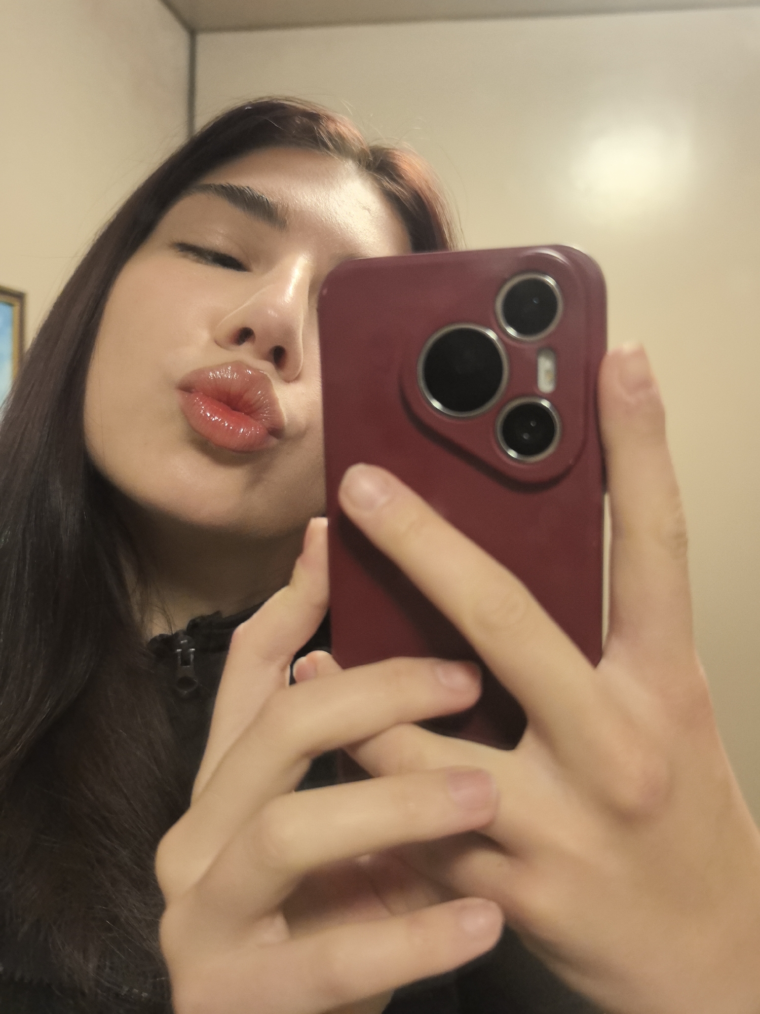 A young woman is taking a selfie indoors, likely in a bathroom, holding a smartphone with a burgundy-colored case. She is making a kissing face. Her dark hair is visible on the left side of the frame. The wall behind her is a light beige color. The location is not identifiable as Stara Zagora, Bulgaria from the visual content provided.