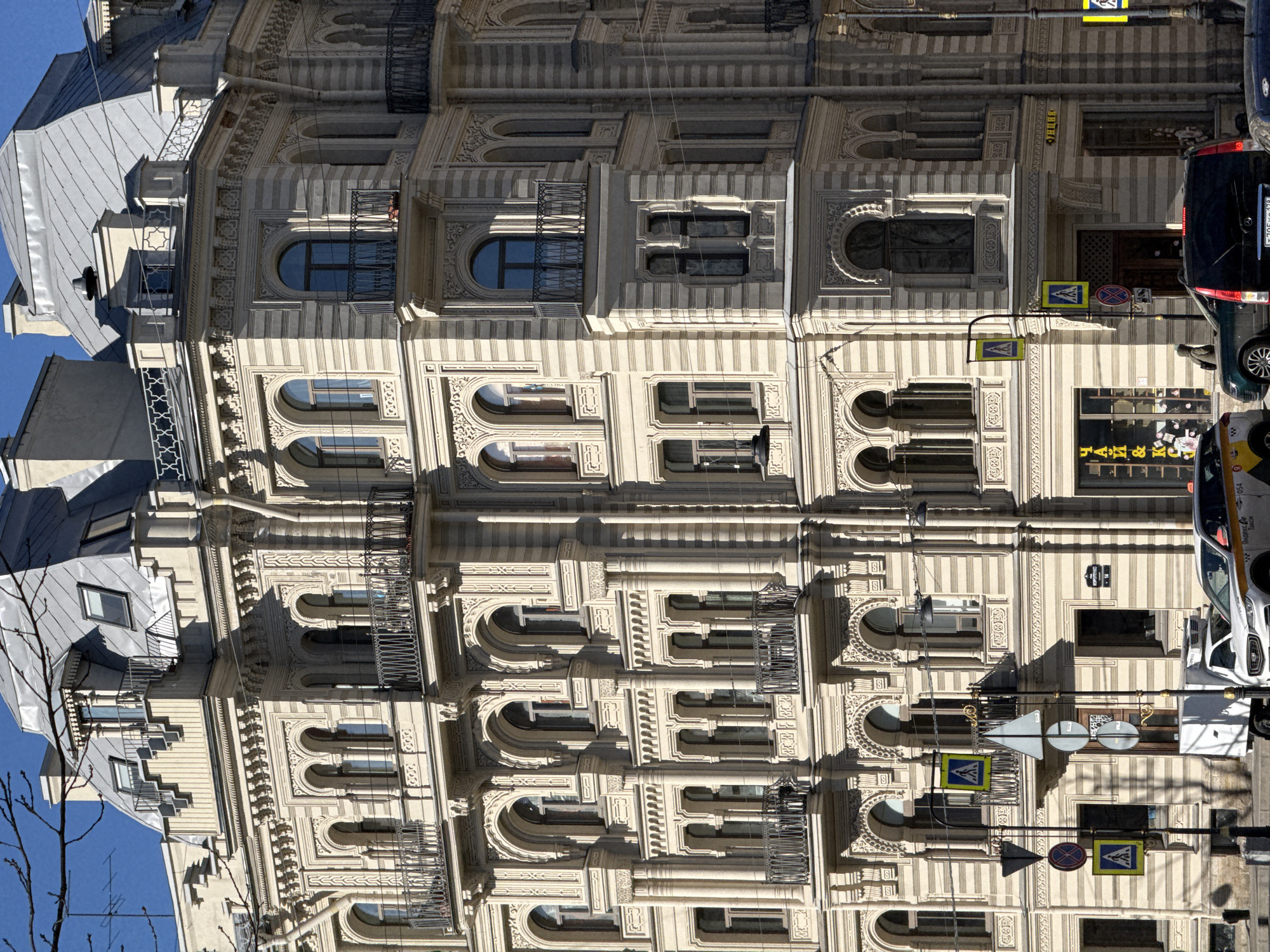 The image shows a building in Saint Petersburg, Russia, specifically the facade of a building at Nevsky Prospect. The architecture features ornate detailing, multiple arched windows, and balconies. The building appears to be several stories tall. Street signs and traffic poles are attached to the building. Several vehicles, including a black SUV, a yellow taxi, and a white van, are parked on the street alongside the building. A bare tree branch is visible in the bottom left corner of the image.