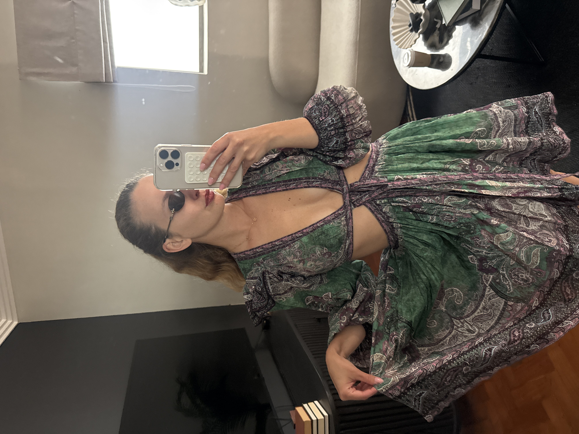 A fair-skinned woman is seen in a full-length, sideways-rotated shot, seemingly taking a selfie in a room with neutral wall paint. She is wearing sunglasses and a patterned, green and purple dress that features cutouts around the midriff. She is holding a phone with a white case.
Behind her, in the reflected background, is a window with a partially closed blind, a light-colored sofa, and a circular black table. A section of a large, black television is visible, and a stack of books is positioned near the bottom of the frame. The scene lacks specific details to determine the exact location, but it is not possible to ascertain whether it is in Cape Town, South Africa.