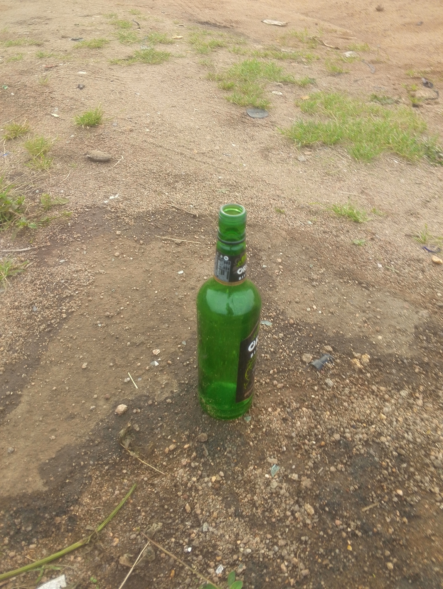 The image shows a green glass bottle lying on the ground in an outdoor area that appears to be a dirt road with patches of dry grass and small pebbles. The bottle has a black label with some text visible, including the word "ORIGINAL". The ground is uneven and appears to be somewhat degraded, possibly due to foot traffic or environmental conditions. The lighting suggests it is daytime. The scene is devoid of any human presence or activity. Based on the provided context, this scene is located in Jos, Nigeria.