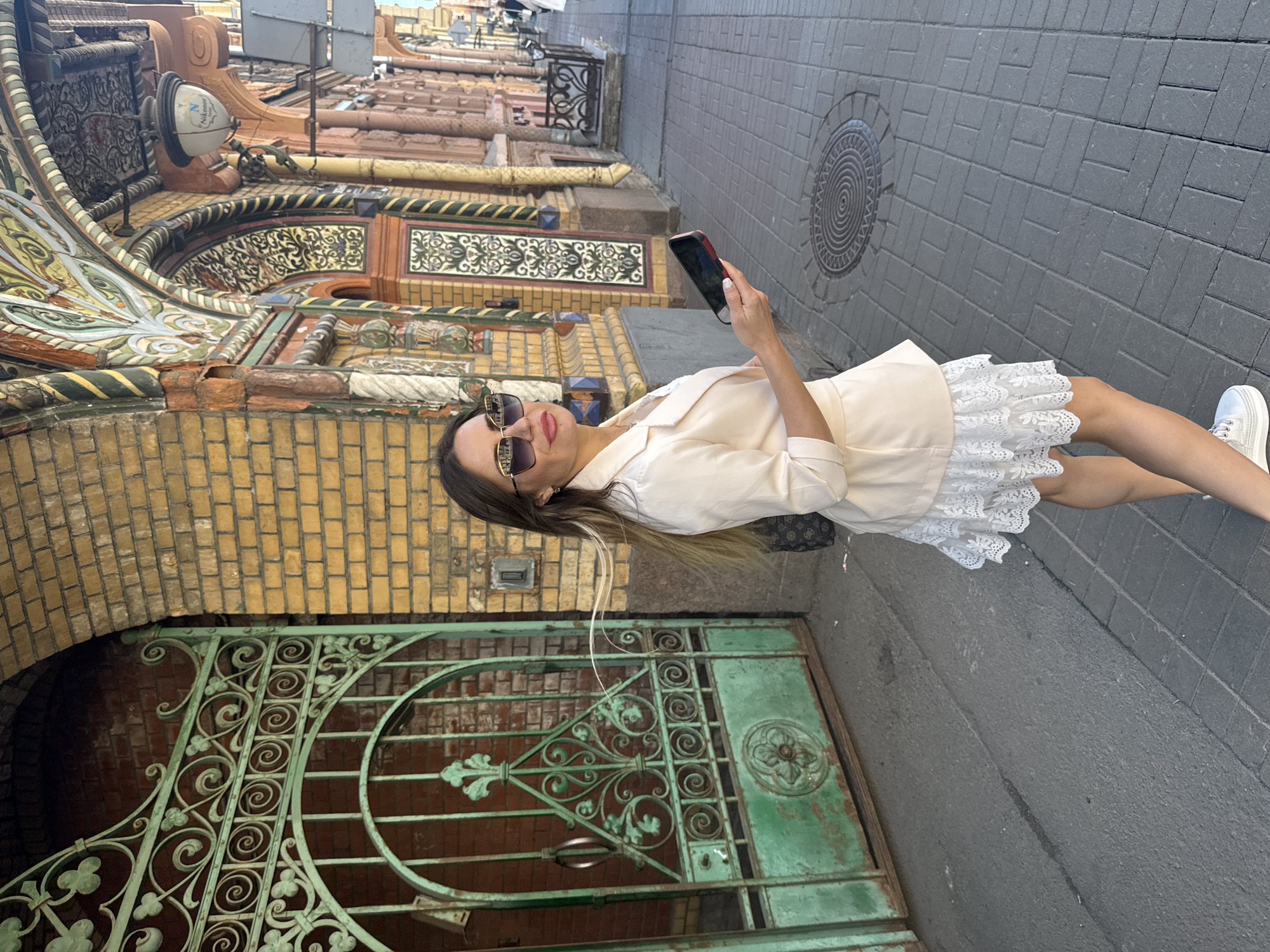 A woman stands on a cobblestone sidewalk, holding a smartphone. She is wearing a light-colored jacket and a white lace skirt. The sidewalk is paved with grey rectangular stones, and a circular metal grate is visible in the foreground. Behind her, an ornate building facade features yellow brickwork and intricate decorative elements in shades of green, yellow, and red. A decorative green metal gate is also visible. The scene appears to be set on a sunny day, given the lighting. The woman is looking at her phone. The architectural style of the building suggests a historical European city, consistent with the provided location context of Saint Petersburg, Russia.