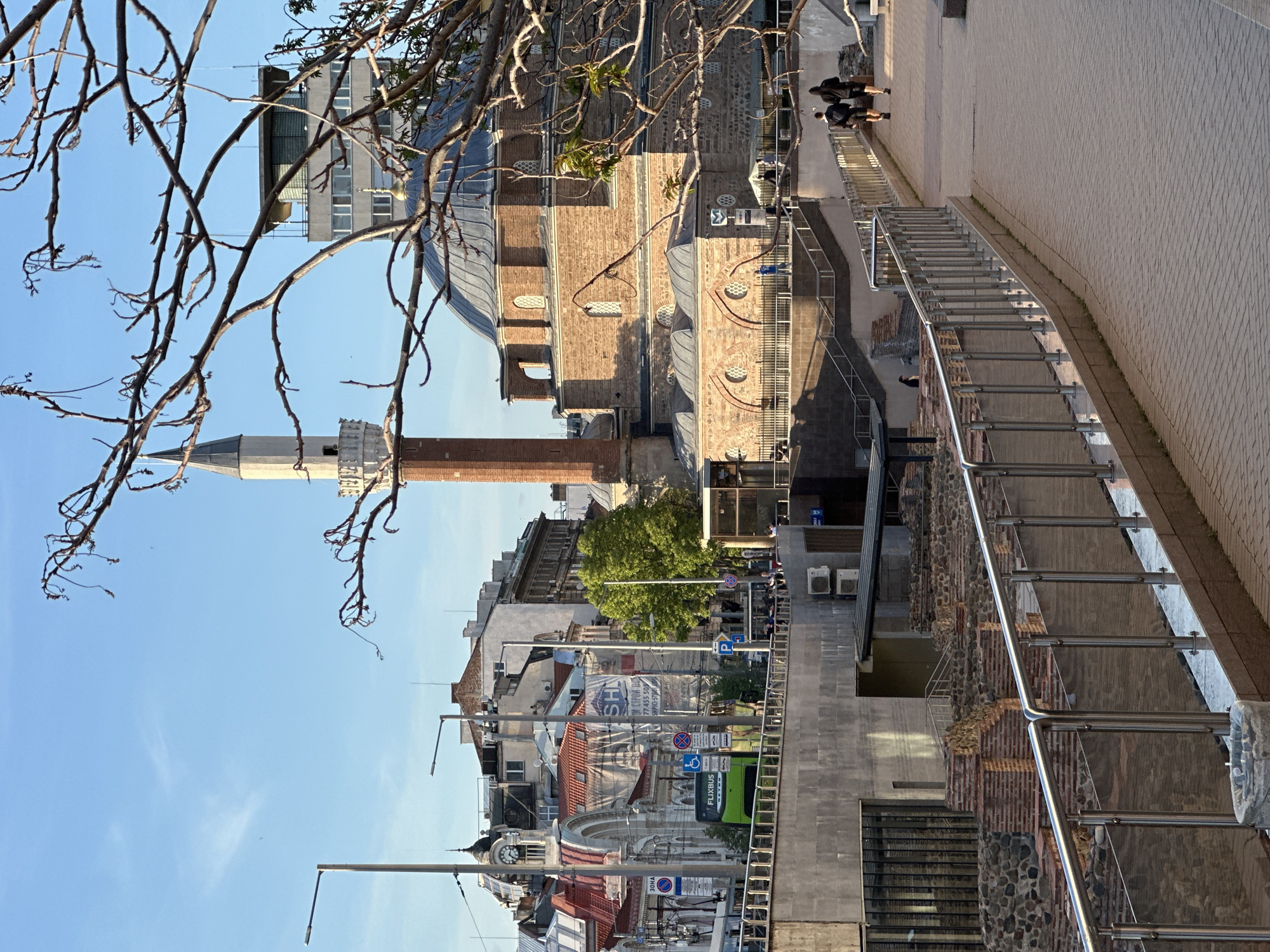 The image shows a streetscape in Sofia, Bulgaria, featuring the Saint George Rotunda. The Rotunda, a reddish-brown brick structure with a dome and a slender minaret, is partially obscured by tree branches in the foreground. A modern staircase with metal railings leads down from a pedestrian walkway alongside the building. The surrounding area includes other buildings of varying heights and architectural styles, a street with pedestrian traffic, streetlights, and a green Flixbus. The sky is blue and clear.