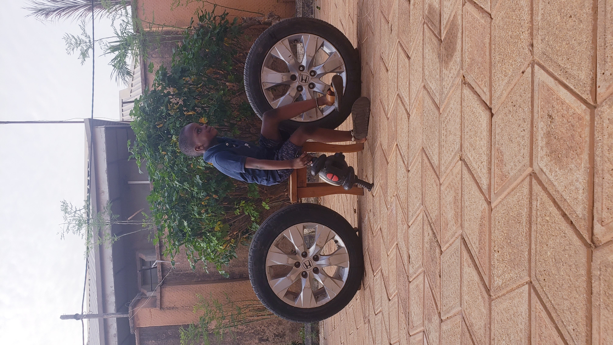An outdoor scene captured in Narayi, Nigeria, features a young boy engaged in play. The boy, wearing a blue polo shirt and dark shorts, is seated on a small, light-brown wooden stool. He is positioned between two large car wheels, which have silver alloy rims and black tires.
The boy is holding a black and red toy power tool, resembling a drill, with both hands. His gaze is directed upwards and to his right, suggesting focus or contemplation as he interacts with his toy. The car wheel rims clearly display the "HONDA" logo on their center caps.
The ground beneath is a paved surface made of reddish-brown tiles arranged in a distinct geometric, diamond-like pattern. In the background, lush green foliage, likely bushes or small trees, provides a natural backdrop. Above the greenery, a light-colored building or wall is visible, topped by what appears to be a grey roof or overhang. The sky above is bright and clear, indicating it is daytime. The lighting suggests a bright day, possibly without direct harsh sunlight.