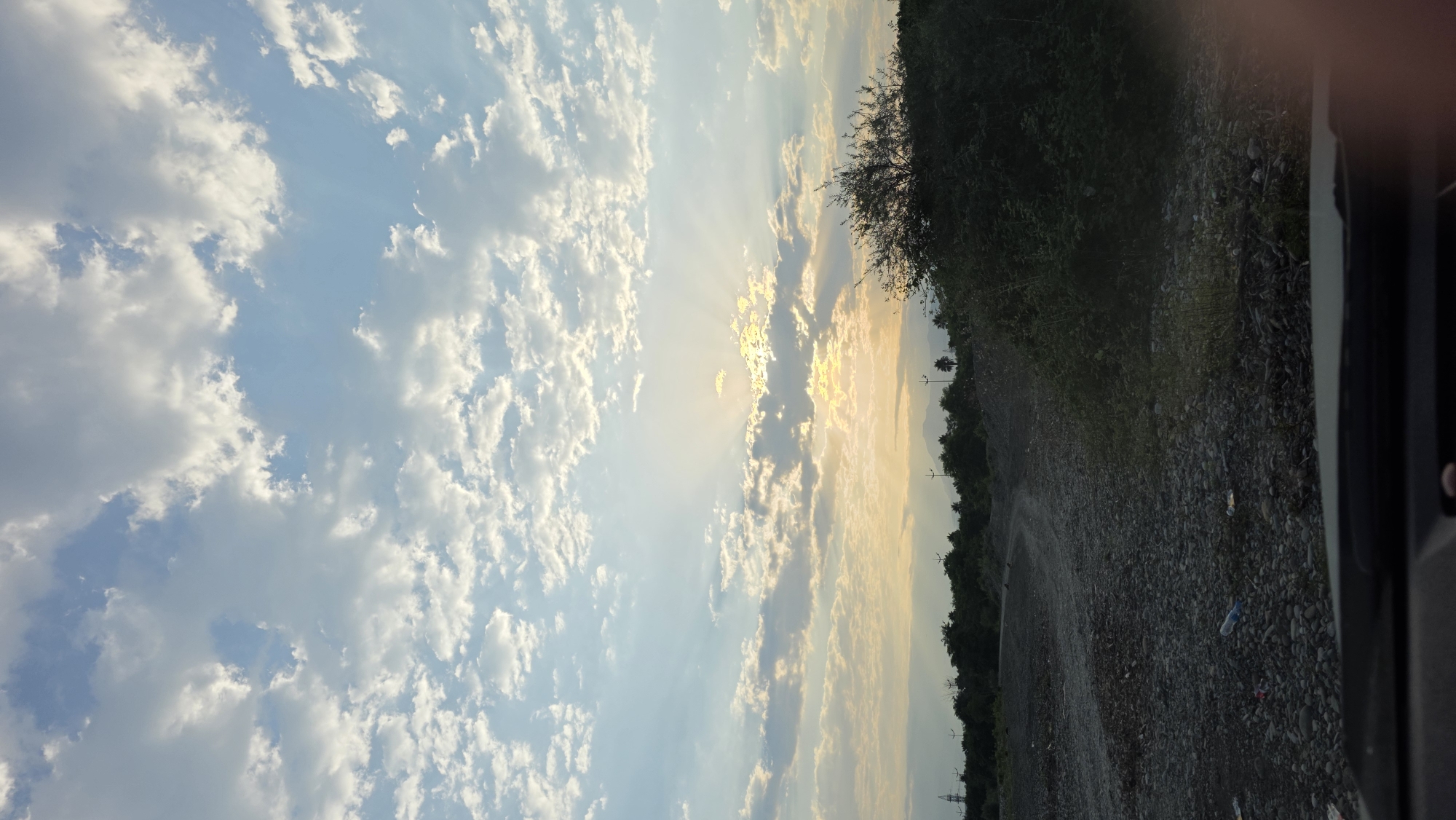 The image captures a scenic view of the sky over the outskirts of Batumi, Georgia. The sky is filled with a mix of fluffy white clouds and patches of clear blue, with the sun peeking through, casting dramatic rays of light. Below the sky, a landscape unfolds with a dirt road winding through a rocky, uneven terrain. To the right, a dense bush is prominent, while a line of trees marks the horizon. In the distance, mountains are visible, and power lines suggest human presence in the vicinity. The foreground shows the dashboard of a vehicle, indicating the photograph was taken from inside a car. The overall impression is one of a natural, somewhat unkempt environment, likely on the outskirts of the city, bathed in the soft light of either sunrise or sunset.