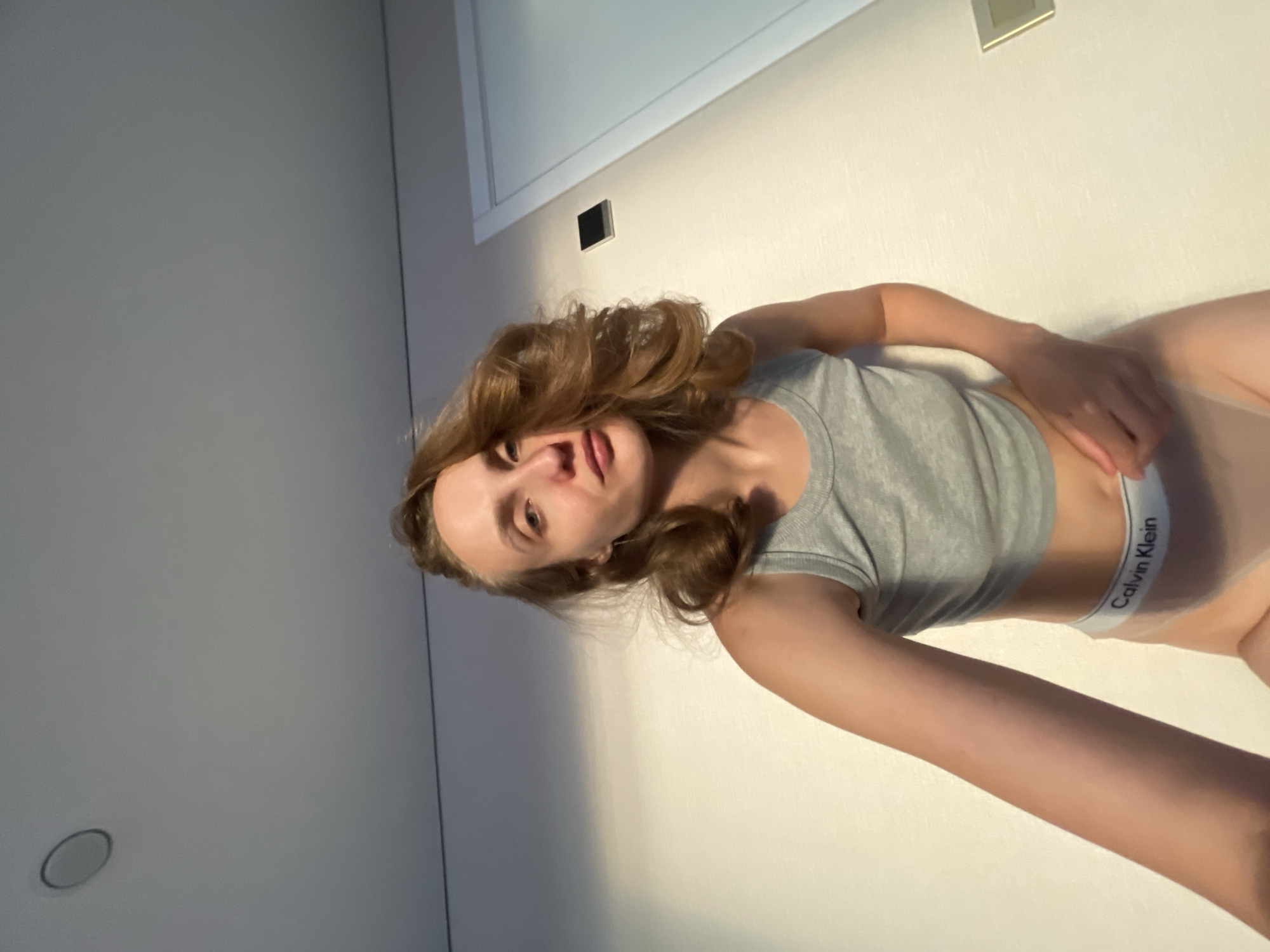 The image shows a young woman lying on her back in what appears to be a room. She is wearing a grey tank top and white Calvin Klein underwear. Her hair is light brown and wavy, and her eyes are open. The lighting suggests it is daytime, with sunlight casting shadows on the wall behind her. There is a subtle hint of a light fixture on the wall above her. The overall impression is a casual, personal moment captured at home. The visible text reads "Calvin Klein" on the waistband of her underwear.