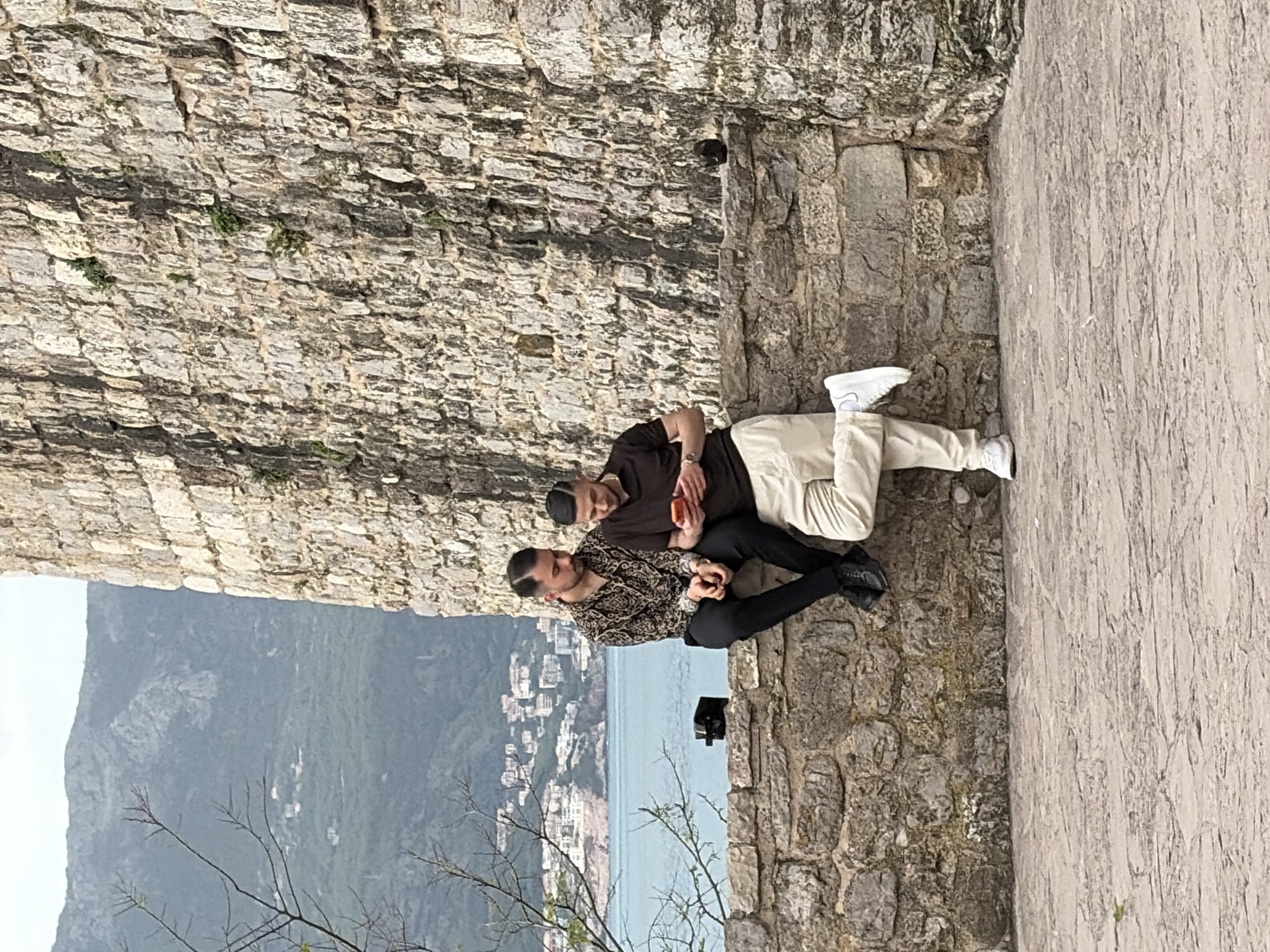 Two men are seated on a stone wall in Budva, Montenegro. One man wears a brown shirt and khaki pants, while the other wears a patterned shirt and black pants. Behind them, a stone wall stretches upwards, and in the background, a view of the city, sea, and mountains is visible. A small tree branch is visible in the foreground. A black object, possibly a bag, sits on the wall near the men.
