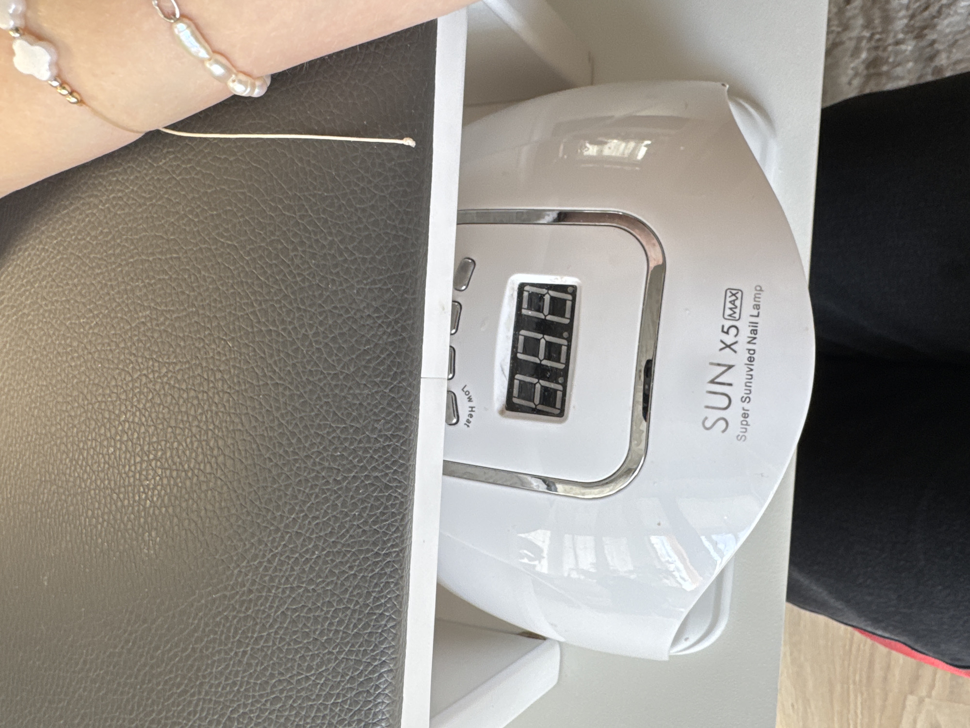 A close-up, overhead shot shows a white nail lamp with a digital display reading "8.8.8." The lamp is labeled "SUN X5 MAX Super Sunuvled Nail Lamp." Above the lamp is a textured, dark gray surface. On the right side of the image, a person's arm is visible, adorned with two pearl bracelets. The background is out of focus but appears to be a domestic indoor setting with a light-colored surface below the lamp. The image does not contain any information related to the city of Perm, Russia.