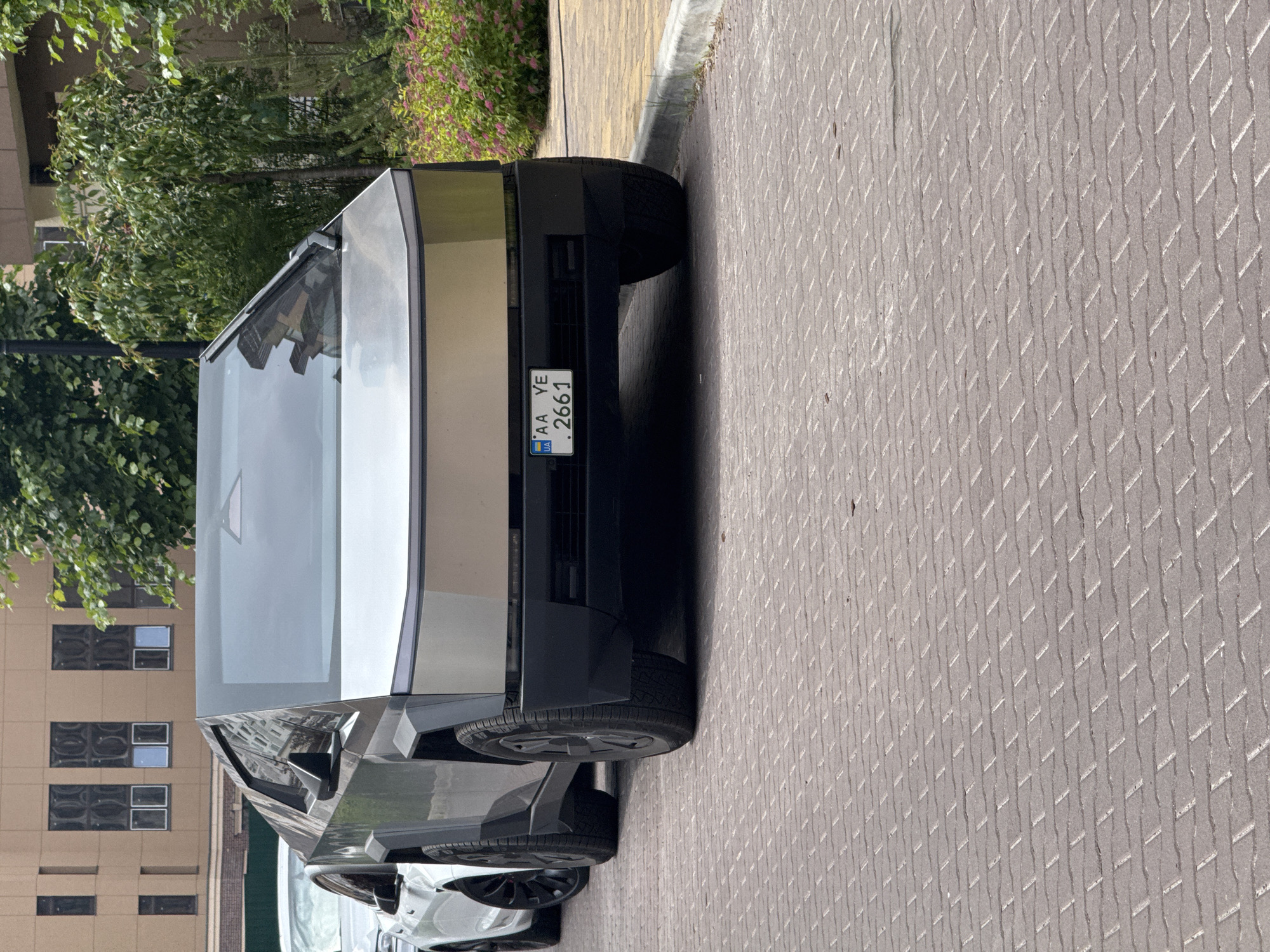 An outdoor daylight photograph captures a silver Tesla Cybertruck parked on a patterned paved surface in Sofiivska Borshchahivka, Ukraine. The Cybertruck, seen from the front, displays its characteristic angular stainless steel body and a black lower fascia. A Ukrainian license plate, "AA YE 2661", is mounted on its front. Behind the Cybertruck, a white car, likely another Tesla, is partially visible, featuring black multi-spoke wheels. The foreground consists of light grey and brown rectangular paving stones laid in an interlocking pattern. To the right of the vehicles, a grey concrete curb borders a landscaped area with green foliage, including trees and bushes with pink flowers. In the background on the left, a multi-story building with a light tan facade and numerous dark-framed windows is visible. The scene is devoid of people.