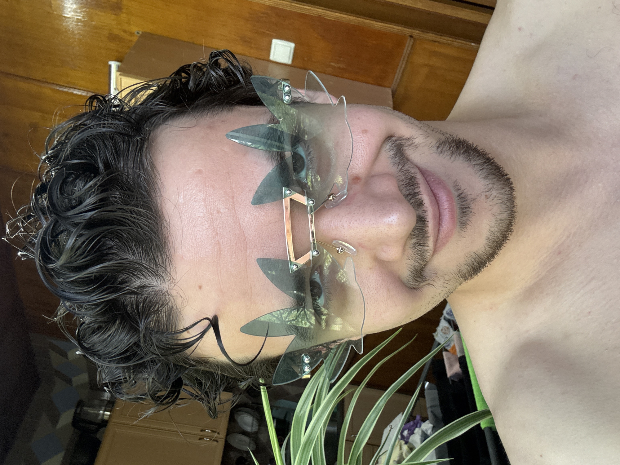 A man is photographed close-up, wearing sunglasses with an unconventional design featuring geometric shapes. His hair is dark and appears wet. He has facial hair, and part of his upper body, specifically his shoulder and neck, are visible. The background includes a wooden surface and a plant with long, green leaves. Given the absence of details confirming the location, I cannot determine if the photo was taken in Tashkent, Uzbekistan.