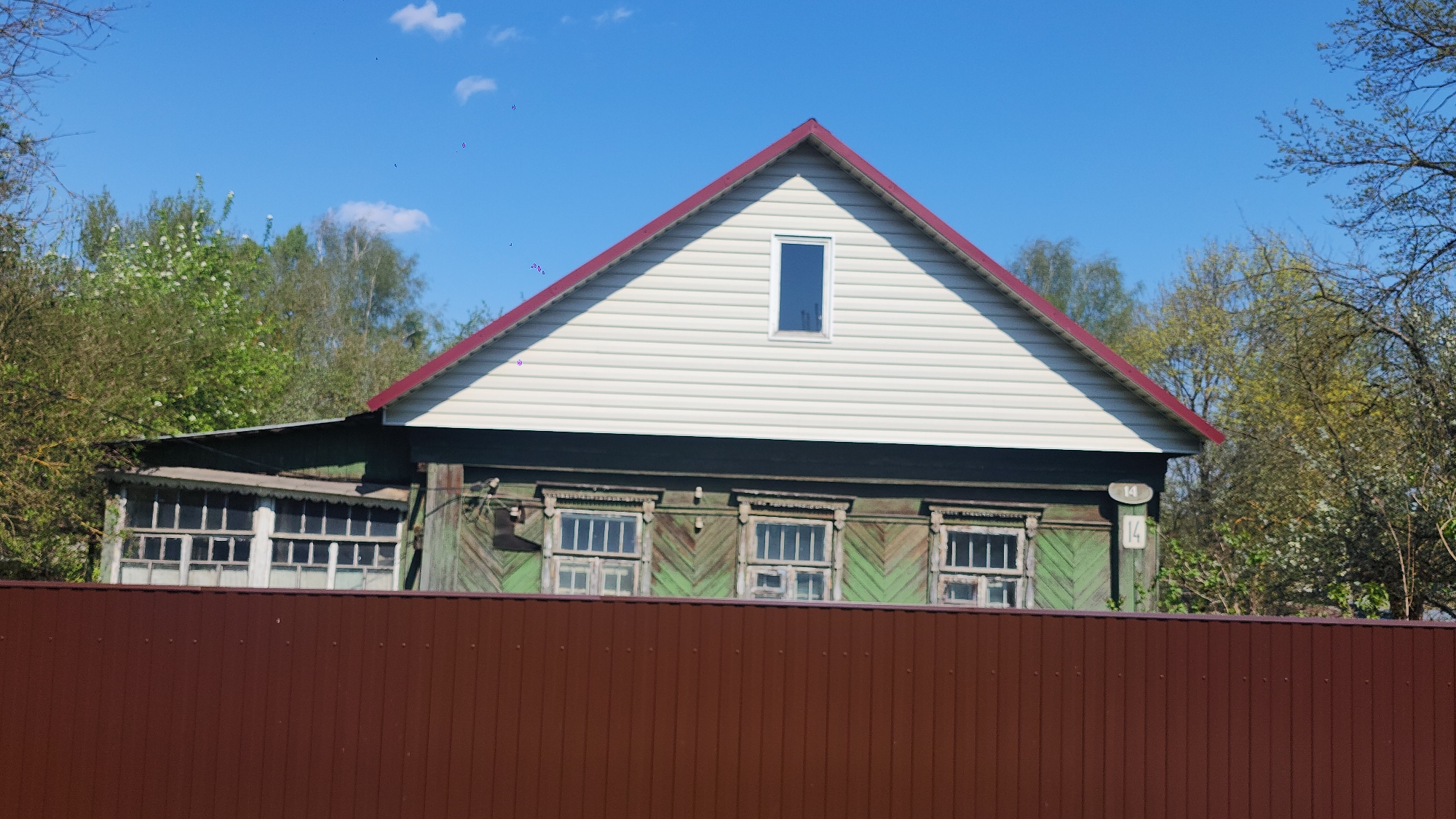 The image shows a weathered wooden house, partially obscured by a brown fence. The house has a red roof and white siding on the gable. The lower portion of the house is painted in shades of green with decorative wooden trim around the windows. A section of the house to the left has a larger window area. Trees with green foliage are visible in the background. The sky is blue with a few scattered clouds. The number "14" is visible on the side of the house. The location is Moscow, Russia.