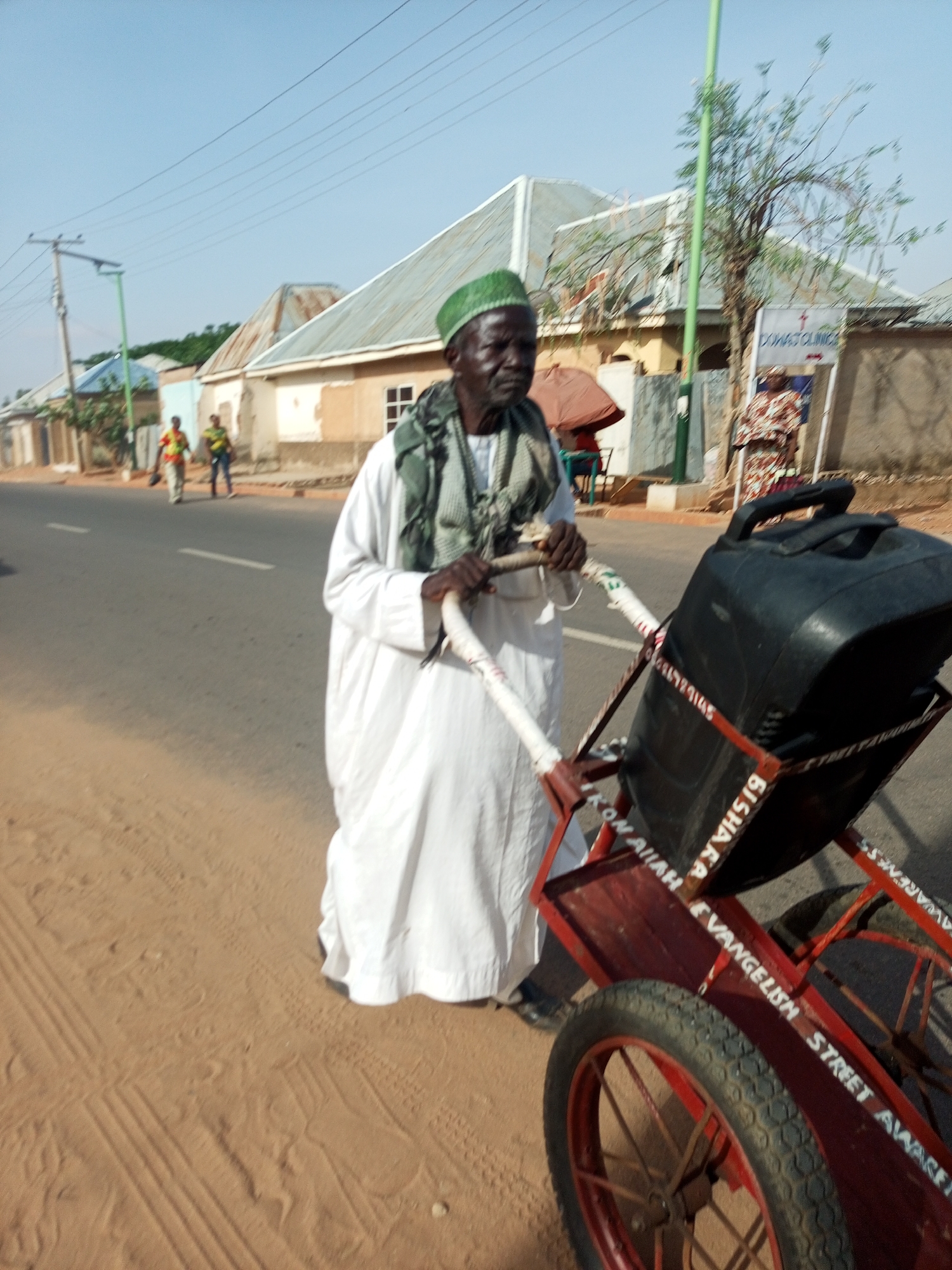 A man in traditional attire, a white robe and green cap, is pulling a red cart with a black object on it along a paved road with sandy shoulders. The cart has writing on it, appearing to say "BISHARA EVANGELISM STREET AWARENESS" and "DIKON ALLAH." In the background are houses and a sign that reads "DOWAS CLINICS." Two people are walking in the distance on the road. The image suggests an urban or semi-urban environment, likely located in Nigeria, as stated.