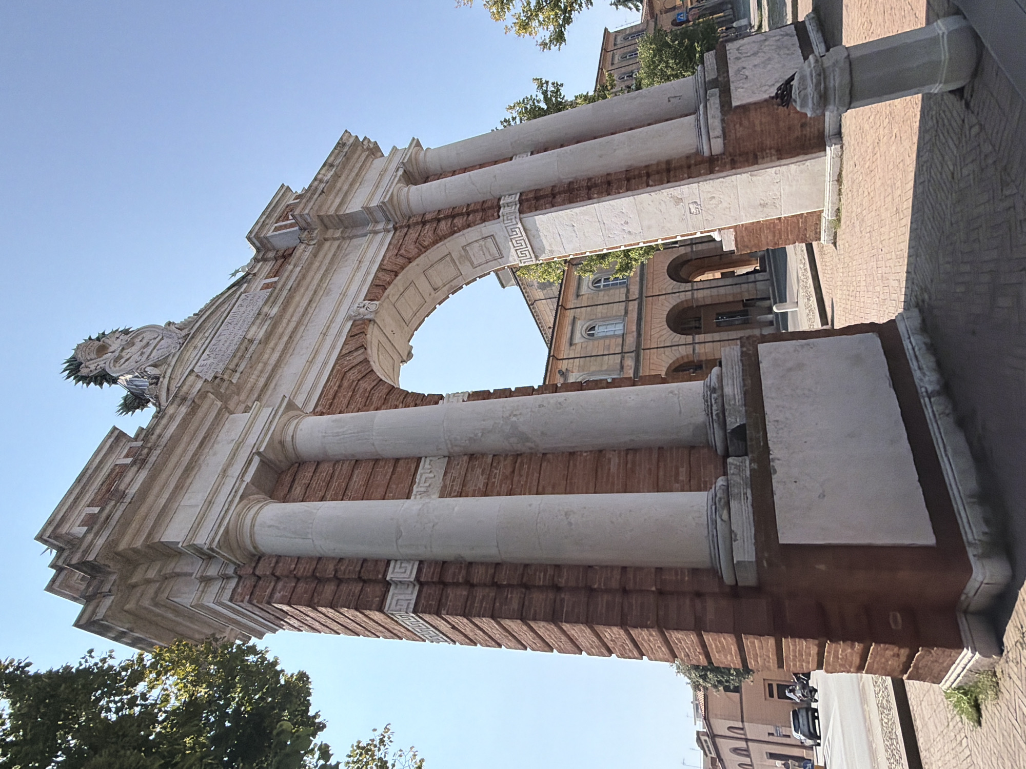 The image shows a low-angle view of the Triumphal Arch of Clement XIII in Santarcangelo di Romagna, Italy. The arch is constructed from red brick and features white marble decorative elements, including columns, cornices, and a sculpted crest at the apex. The inscription on the arch is partially visible. The sky is clear blue. In the background, a building with arched walkways and windows can be seen, along with trees and a street with a car and a scooter. The ground in front of the arch is paved with bricks.
