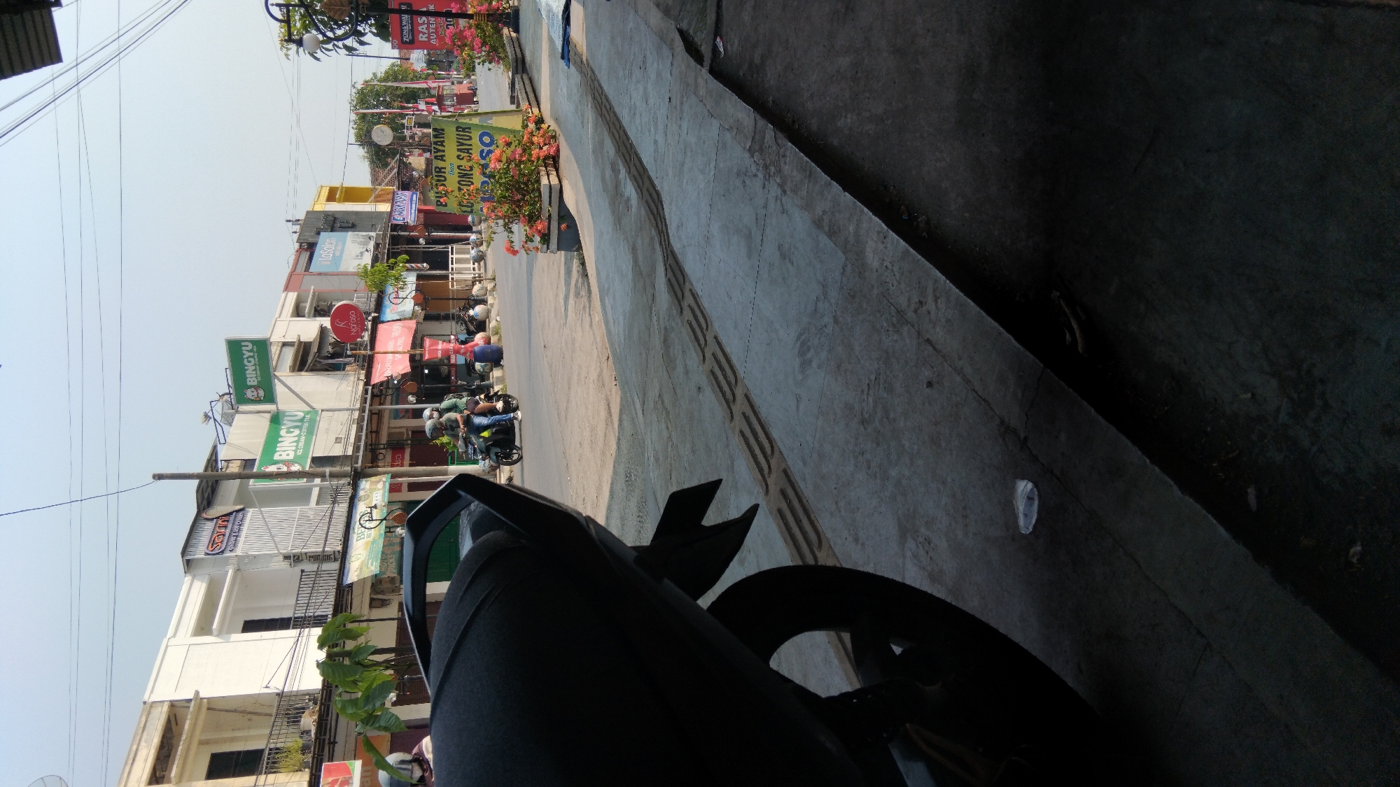 The image depicts a street scene in Batang, Indonesia, likely during daylight hours with clear skies. In the foreground, the rear section of a dark-colored motorcycle is visible, partially obscuring the left side of the frame. The motorcycle's tire and part of its bodywork are in focus. Adjacent to the motorcycle is a concrete sidewalk featuring raised tactile paving, indicating it is likely a pedestrian pathway. A few pieces of litter, including a clear plastic cup, are scattered on the sidewalk.
The background reveals a street with several buildings lining one side. These buildings appear to be commercial establishments, with various signs displayed. Prominent signs include "BINGYU ICE CREAM COFFEE TEA" in green and white, and another "BINGYU" sign. Other visible signs are partially obscured or unreadable due to distance and resolution. A few people are present on the street. Two individuals are riding a motorcycle in the middle ground, heading away from the camera. They are wearing helmets. On the sidewalk in front of the buildings, a few more individuals appear to be engaged in activities related to their businesses or the street. One person is standing near a display of flowers, and another is near what appears to be a food stall or cart. Various potted plants and flowers are arranged along the sidewalk in front of the establishments, adding some color to the scene. Power lines crisscross the sky above the street. The overall impression is a typical urban street scene in a commercial area.