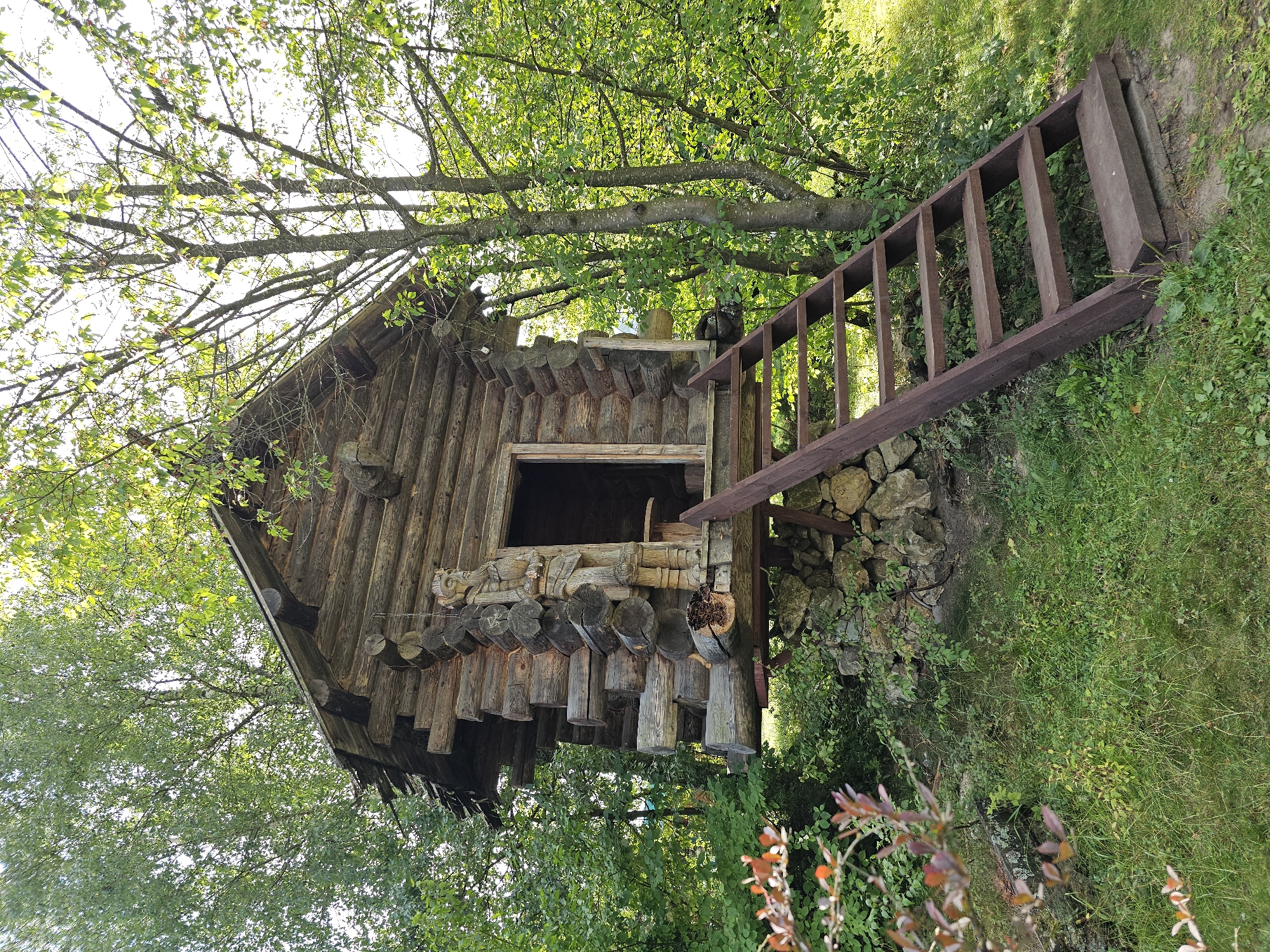 The image depicts a rustic, wooden log cabin elevated on a stone foundation, with a wooden staircase leading up to its entrance. The cabin has a steeply pitched roof and is situated amidst lush green foliage and trees, suggesting a natural, possibly rural, setting. The architectural style of the cabin, constructed from logs and featuring a carved wooden figure next to the doorway, hints at traditional Russian folk design. The ground surrounding the cabin is covered in grass and some brush. The overall scene evokes a sense of a secluded, perhaps historical, dwelling in a natural environment. The location is identified as the city of Ulitkino, Russia.