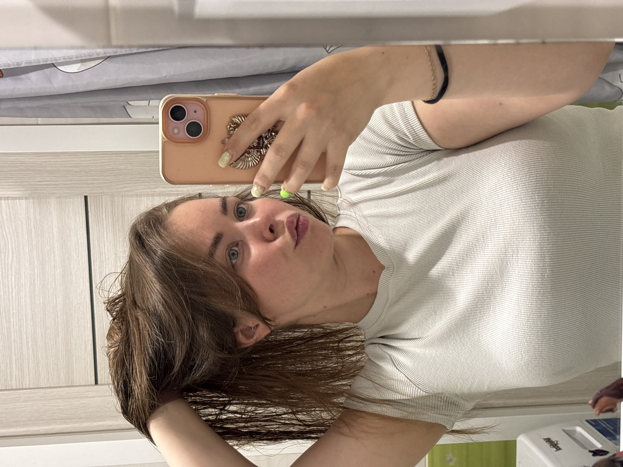 The image shows a woman with light skin, brown hair, and blue eyes taking a selfie in what appears to be a bathroom. She is holding a peach-colored iPhone with a decorative case in her right hand. She is wearing a light-colored ribbed shirt. Her left hand is touching her wet hair. She has a black and gold bracelet on her left wrist and a ring on her right hand. Her fingernails are painted a light glittery color with one nail painted bright green. The background includes a mirrored wall above a cabinet door, and a glimpse of a curtain above her head. The location is not discernible as being in Izhevsk, Russia.