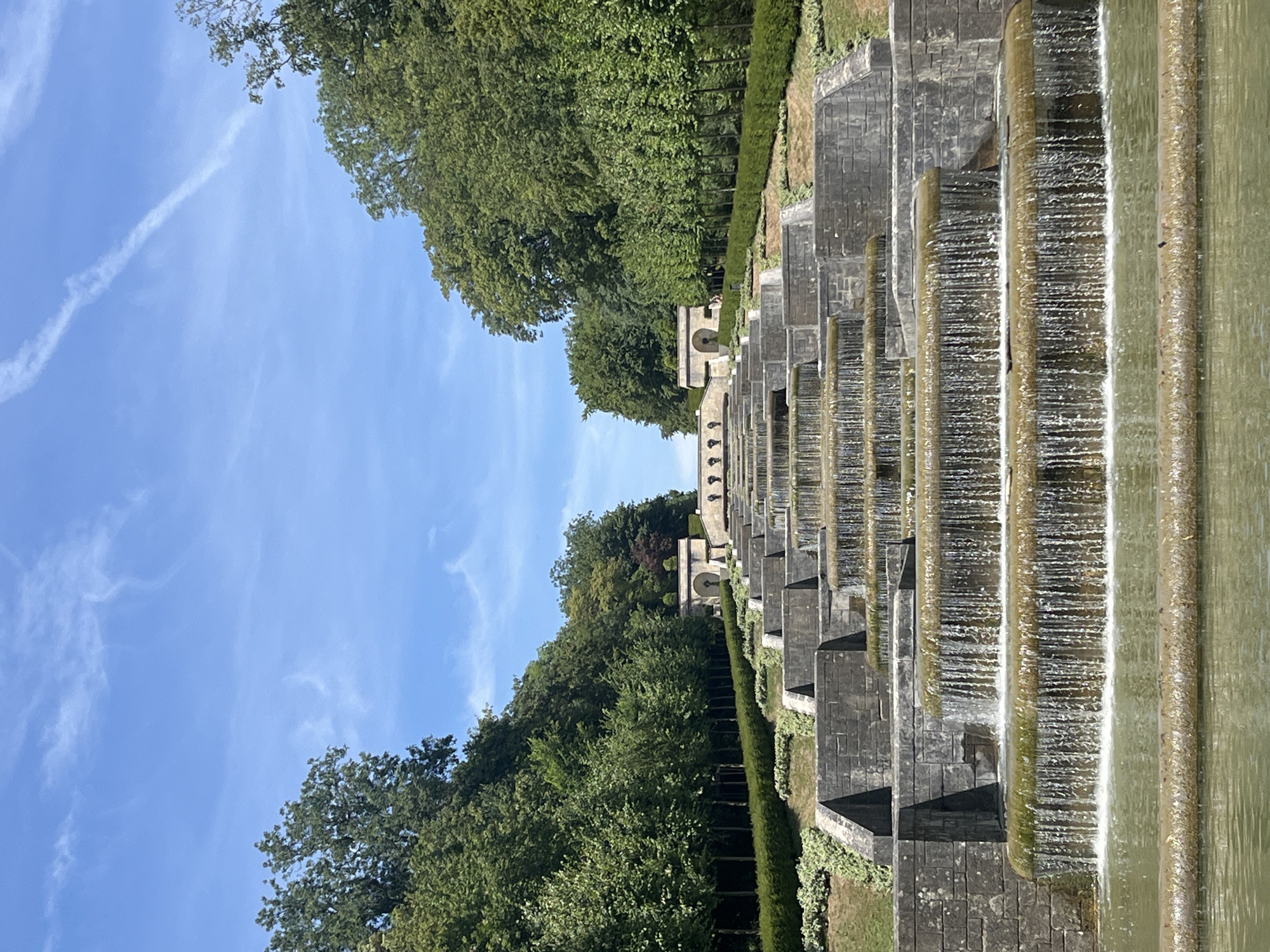 The image shows the Cascade de la Cascade, located in Antony, France. The cascading fountain is constructed from stone and features multiple tiers of flowing water. Lush green trees line the upper sides of the cascading structure, and a clear blue sky with wispy clouds forms the backdrop. The fountain is set within a manicured landscape with hedges and grassy slopes. A pool of water at the bottom of the fountain reflects the surrounding scene.