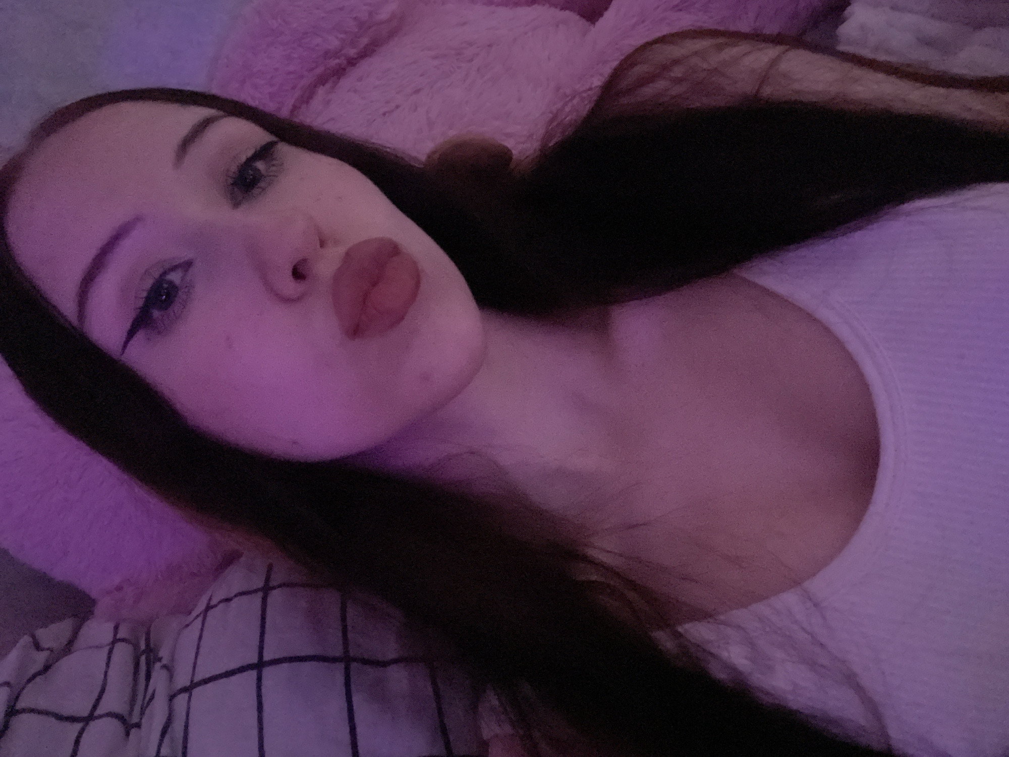 A young woman with long dark hair is looking at the camera with her lips pursed. She is wearing a white tank top. She has winged eyeliner and full lips. She is leaning on a plush pink object. The lighting is dim and purple. The background is blurred, but appears to include a plaid pillow. The scene appears to be indoors. There is no indication of the location being Votkinsk, Russia.