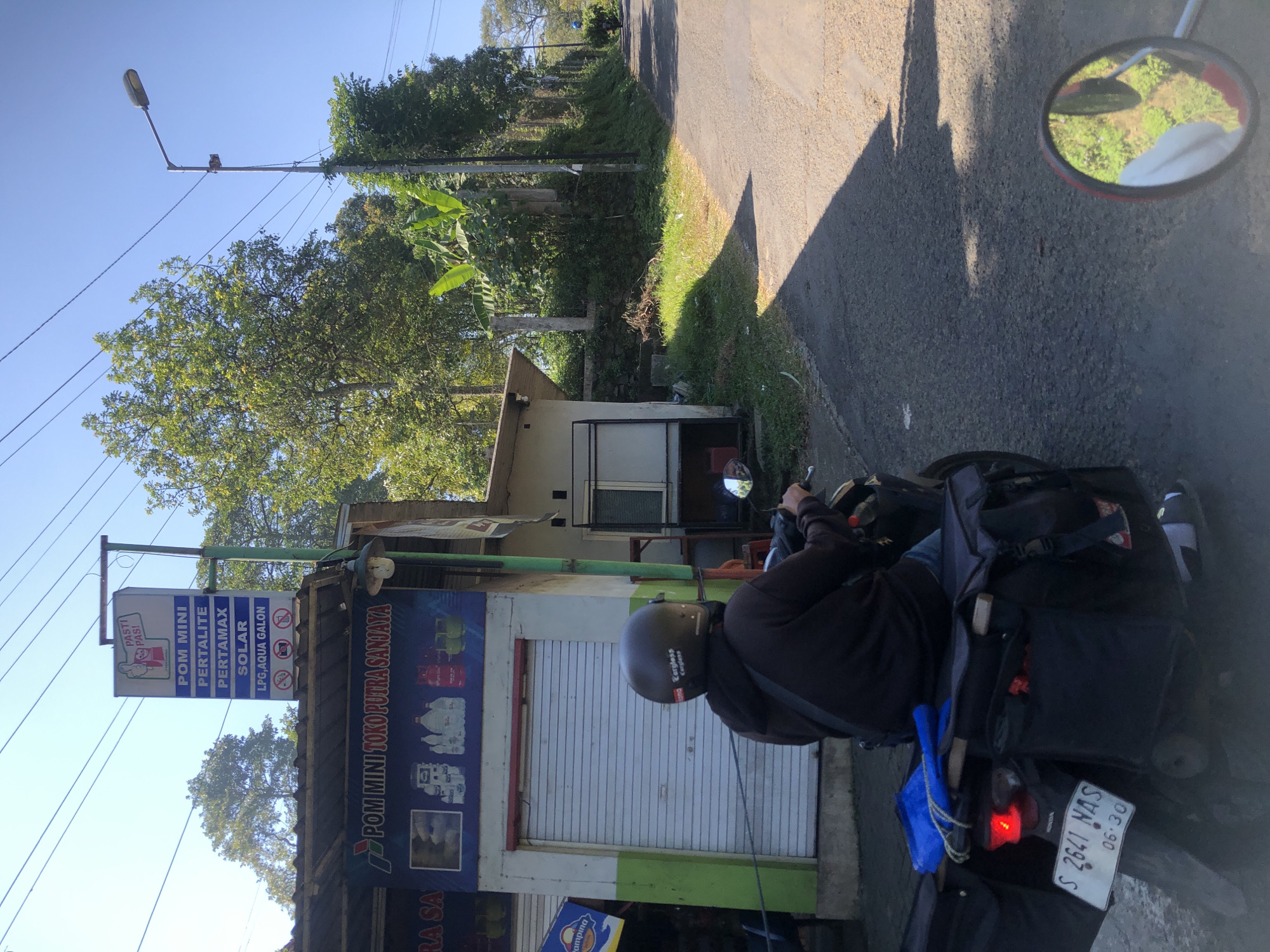 The image depicts a street scene in Trawas, Indonesia, on a bright, sunny day. In the foreground, a person wearing a dark hoodie and a helmet is riding a motorcycle. The motorcycle is equipped with black saddlebags on either side and a blue bag on top. The license plate of the motorcycle reads "S 2641 NAS 06.30". To the left of the motorcycle, there is a small roadside shop with a white and green facade. A sign above the shop reads "POM MINI TOKO PUTRA SANJAYA" and displays images of various products. Further to the left, another sign lists fuel types: "POM MINI", "PERTALITE", "PERTAMAX", "SOLAR", and "LPG, AQUA GALON", along with symbols indicating no smoking, no photography, and no pets.
The road is paved and slopes upwards to the right. A street light is visible on the right side of the road, near a cluster of trees. The environment is lush with green foliage, indicating a semi-rural or suburban setting. In the background, another motorcycle is seen in the distance, also heading uphill. The overall impression is of a typical day on a road in a town or village in Indonesia.