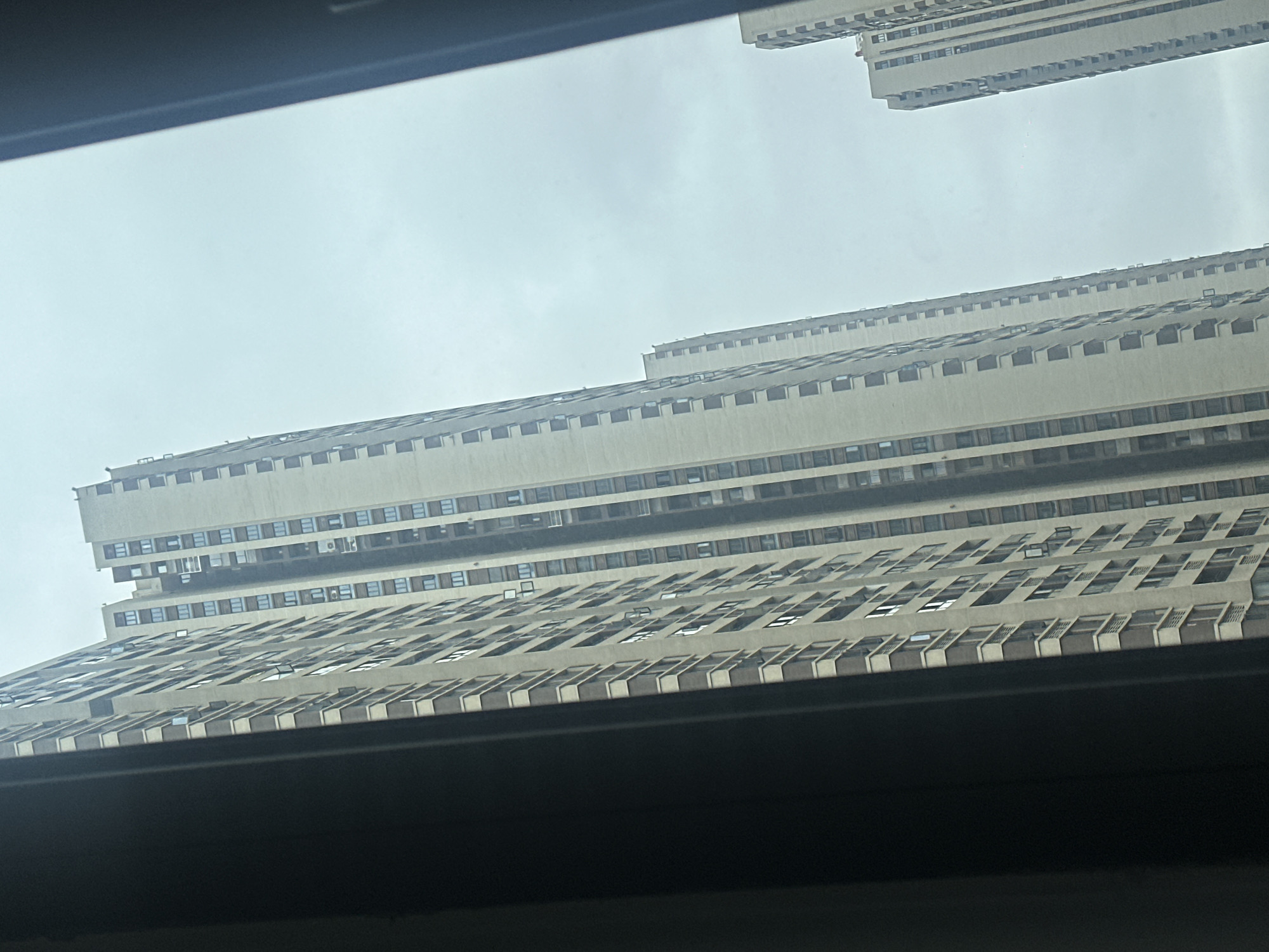 The image displays an upward-angled view of several densely packed high-rise residential buildings within the urban environment of Chongqing High-tech Zone, China. The structures are characterized by light beige or cream-colored facades, prominently accented with dark brown horizontal and vertical trim around numerous windows and balconies. Multiple windows are visible, some appearing partially open, and various exterior air conditioning units are affixed to the building surfaces. The scene is framed on the left and right by dark, opaque structural elements, likely part of a window or door frame, partially obscuring the view. Above the buildings, the sky is a uniform, light grey-blue, consistent with an overcast or hazy atmospheric condition. No individuals, animals, or explicit actions are discernible within the frame.