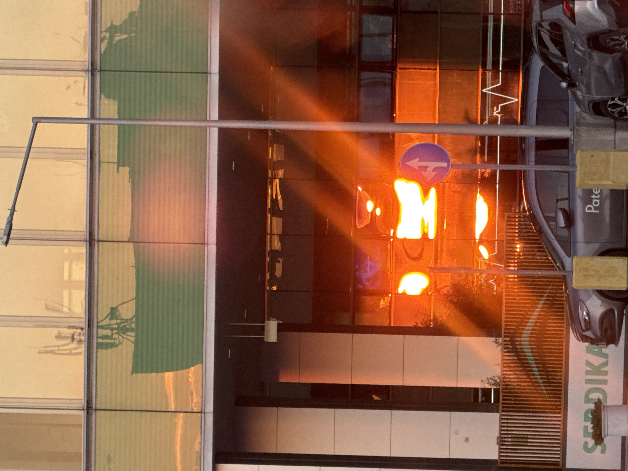 The image captures a building facade at sunset, with sunlight reflecting off the glass windows and casting long shadows. The scene appears to be urban, with hints of a city environment suggested by the presence of a car parked on the right side of the frame. The car is silver and partially visible, with its rear end towards the viewer. A traffic sign indicating a left turn is also visible, further confirming an urban setting.
The building itself is modern, characterized by large glass panels and a metallic structure. The sunlight creates dramatic streaks of orange and yellow across the facade, highlighting the architectural details and the reflective surfaces. The time of day is clearly sunset due to the warm, directional light and the long shadows. There are no people visible in the frame.
Notable details include the text "Pate" and "SERDIKA" printed on a sign or part of the building's branding. "SERDIKA" is a historical name for Sofia, the capital of Bulgaria, providing a strong location cue. The overall mood is serene and slightly dramatic, owing to the intense colors of the sunset and the interplay of light and shadow. The weather appears clear, as the sunlight is strong and unobstructed. The image is oriented vertically, suggesting it might be a detail from a larger scene or captured with a phone.