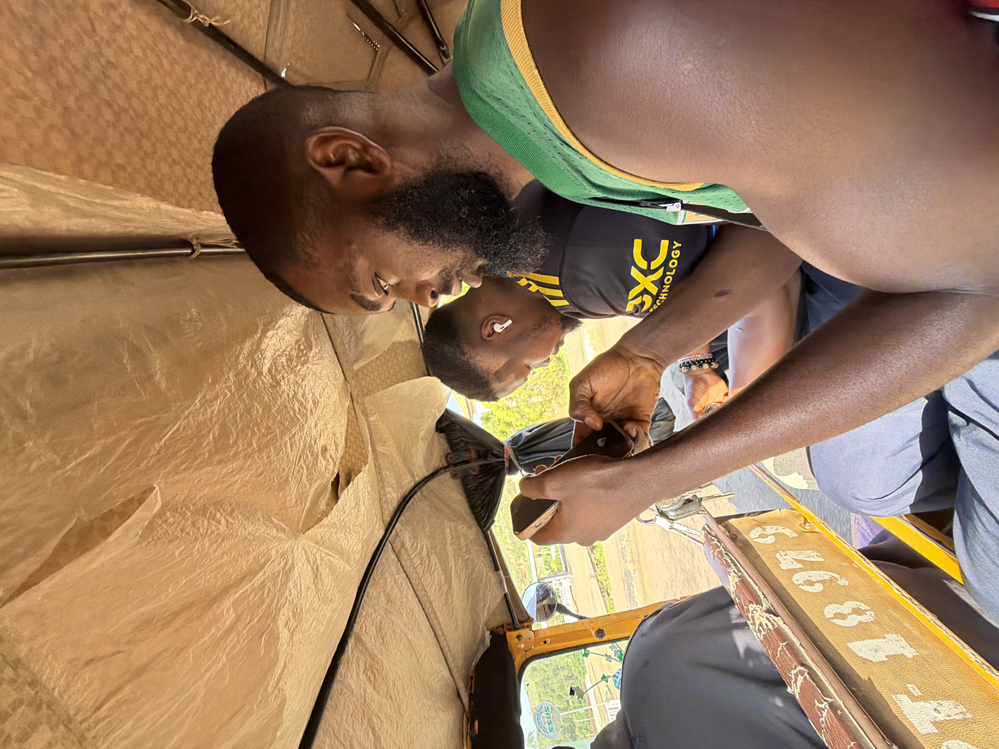 The image shows the interior of a motorized rickshaw in Gura Top, Nigeria. Two men are visible. In the foreground, a man with a beard is looking at his phone while seated on the right. His right arm is extended, and he is holding two mobile phones. He is wearing a green and yellow sleeveless jersey. Behind him and to the left, another man is also looking at his phone. He is wearing a black t-shirt with yellow stripes and the text "EXC TECHNOLOGY" on it. He also has white earbuds in his ears. The rickshaw's interior is lined with a tan-colored fabric, and metal bars support the roof. Outside the rickshaw, through the open side, a blurred background of trees and a dirt road is visible. A license plate with the partial number "T-18979" is attached to the side of the rickshaw.