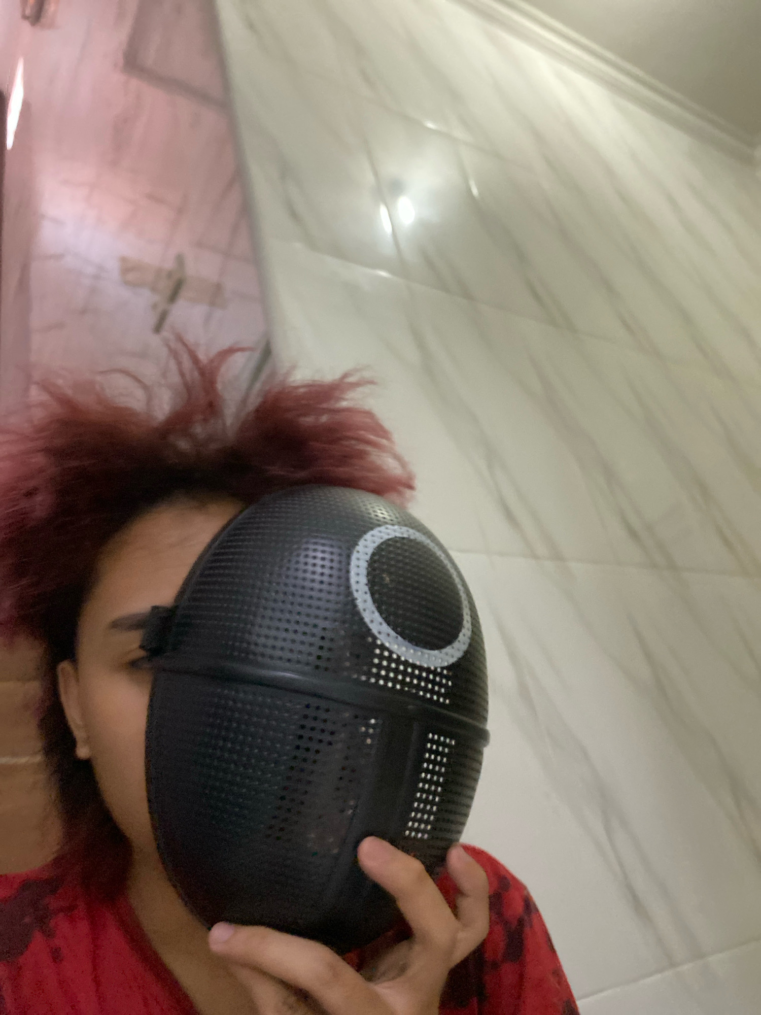 The image depicts a person, partially obscured by a black mask with a circular design on it, holding the mask up to their face. The person has reddish, spiky hair and is wearing a red garment with a dark pattern. The background appears to be a bathroom with white, textured tiles on the wall and ceiling. The lighting suggests an indoor setting. There is no visible indication of the location being in Klurak, Indonesia.