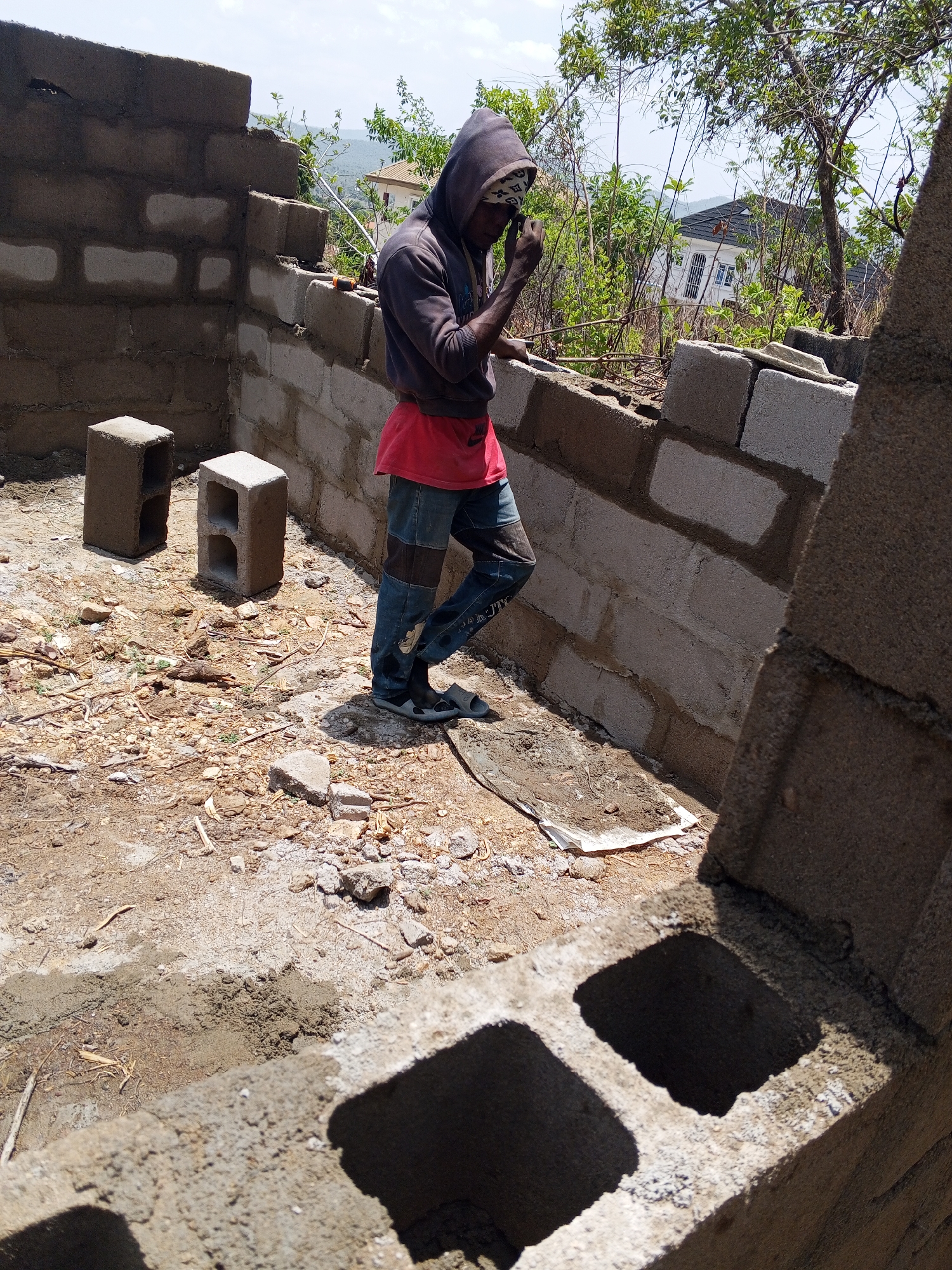 A man is constructing a cinder block wall on a construction site in Lokoja, Nigeria. He is wearing a hooded sweatshirt, a red t-shirt, patchwork jeans, and sandals. The man has a bandana over his head and appears to be adjusting it. Two cinder blocks sit on the ground near a completed section of the wall. The scene is outdoors with a view of trees and a house in the background. The ground is dirt and scattered with debris.